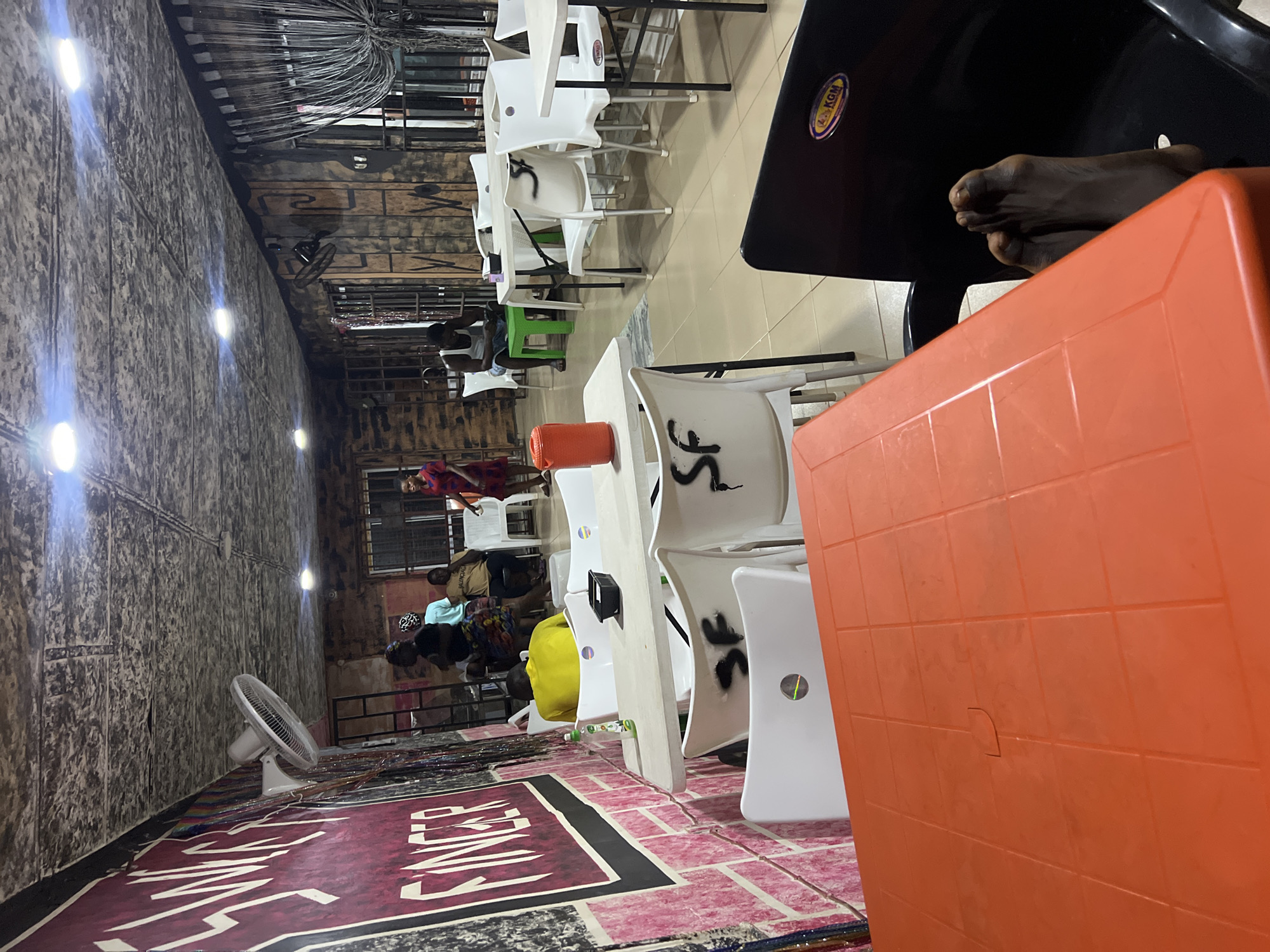 This image depicts the interior of a casual eatery or restaurant, likely at night given the reliance on artificial lighting. The setting appears to be in Obosi, Nigeria.
The foreground features an orange plastic table positioned prominently, angled towards the viewer. Beside it, a black plastic chair with a "KGM" sticker on its back is partially visible, with a person's bare feet resting on it. Several white plastic chairs, some marked with black graffiti-like letters such as "SF," are arranged around white tables, extending into the midground.
In the midground, several people are seated at various tables. One individual in a red top is visible further back, and another person wearing a light blue top and possibly a yellow jacket or bag can be seen. A white standing fan is placed on the patterned floor, which consists of pinkish tiles and a dark mat or rug. The rug has white lettering, with "SINCE" clearly visible, and a larger, less distinct word beneath it that appears to be "FINGER" or similar.
The background is dominated by a textured wall, possibly brick or concrete, illuminated by several bright, upward-facing artificial lights. Near the ceiling, a dense array of wires or pipes is visible. The overall ambiance is casual and relaxed, with patrons appearing to be conversing or waiting. The lighting from the overhead fixtures and the general setup suggest a local establishment catering to casual dining or socializing.