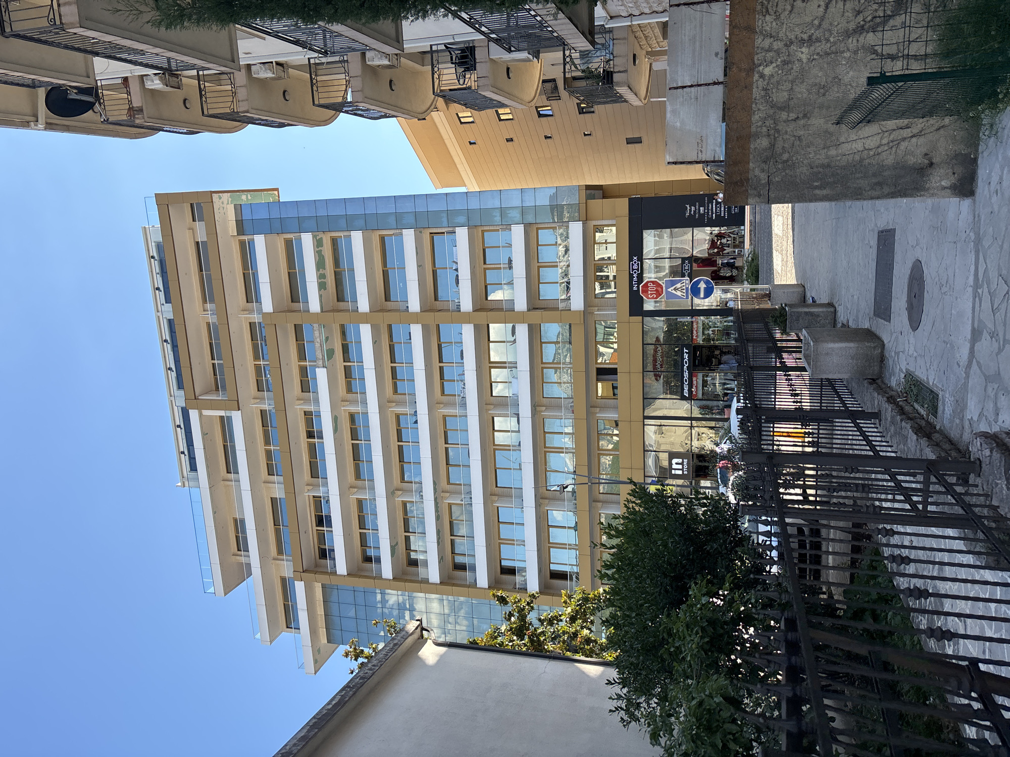 The image captures a view of a modern, multi-story building with a beige and glass facade, situated in the city of Budva, Montenegro. The building has multiple balconies with glass railings and is surrounded by other buildings, one with a similar architectural style and another with a more traditional design featuring balconies with wrought-iron railings. A street with shops at the ground level is visible in front of the modern building. There are several street signs, including a stop sign and directional signs. The sky is clear and blue, suggesting a sunny day. The foreground shows a sidewalk and a black metal fence. The scene appears to be a commercial and residential area in Budva.