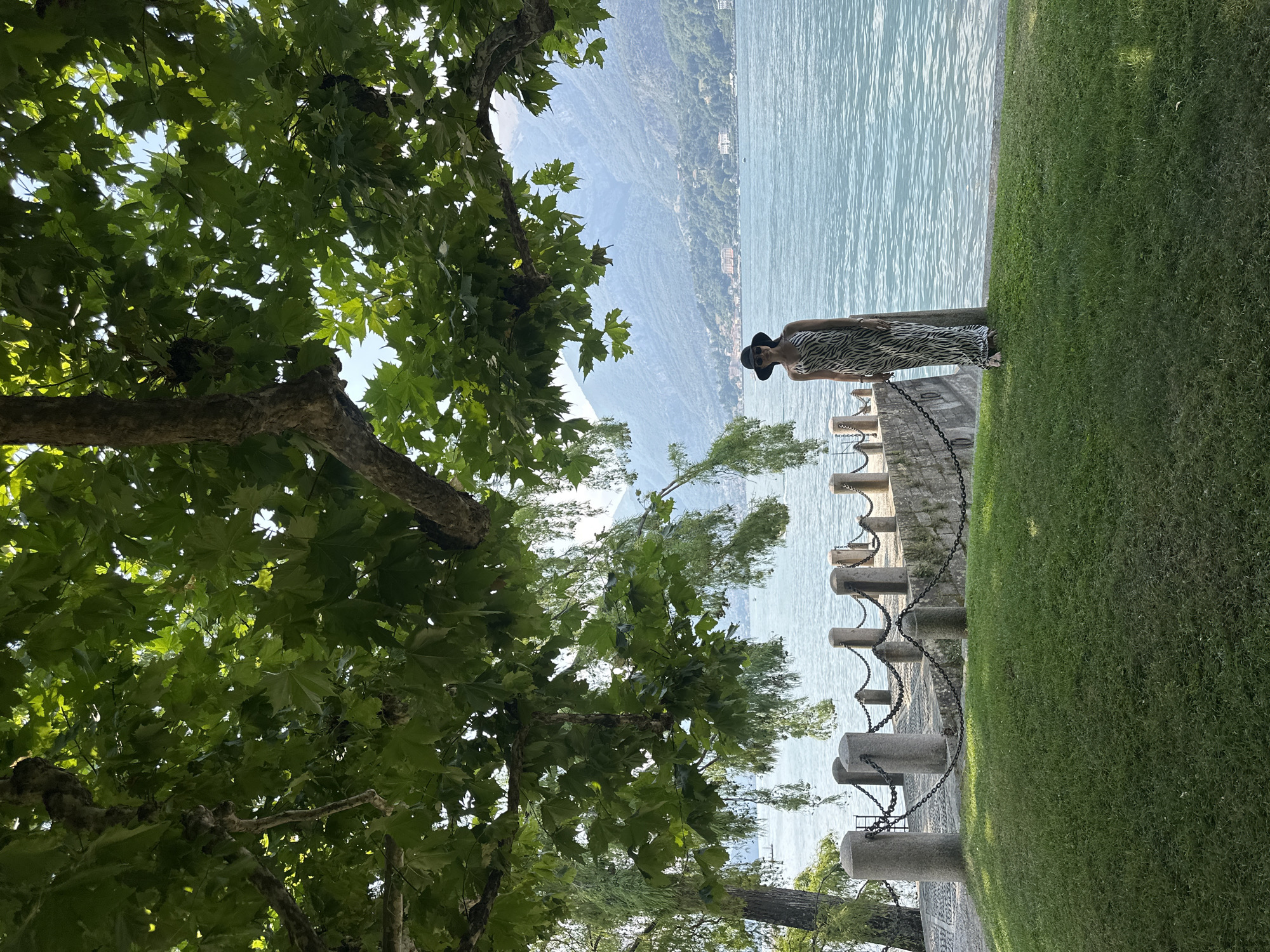 A woman stands on a grassy lawn under the shade of a large tree by a body of water. She is wearing a black hat and a long, black and white zebra-print dress. Behind her, a stone pier with cylindrical bollards and a chain fence extends into the water. The water is a calm blue and reflects the light. In the background, hazy mountains rise above a distant shoreline with buildings. The scene appears to be in a park or lakeside area, possibly in San Giovanni, Italy, given the presence of mountains and water.