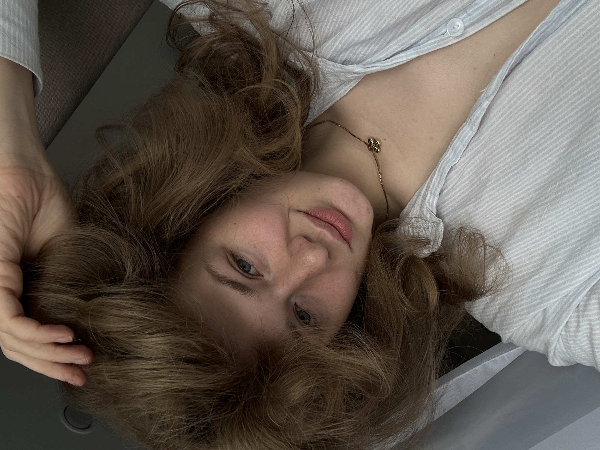 A woman lies on a gray surface, possibly a bed or sofa, indoors. She has light skin, light eyes, and brown curly hair. She wears a light-blue striped shirt that is slightly unbuttoned, revealing a gold necklace with a clover pendant. Her left hand is raised and positioned near her head in the hair. The location is unknown and there is no evidence to suggest the location of Kazan, Russia.