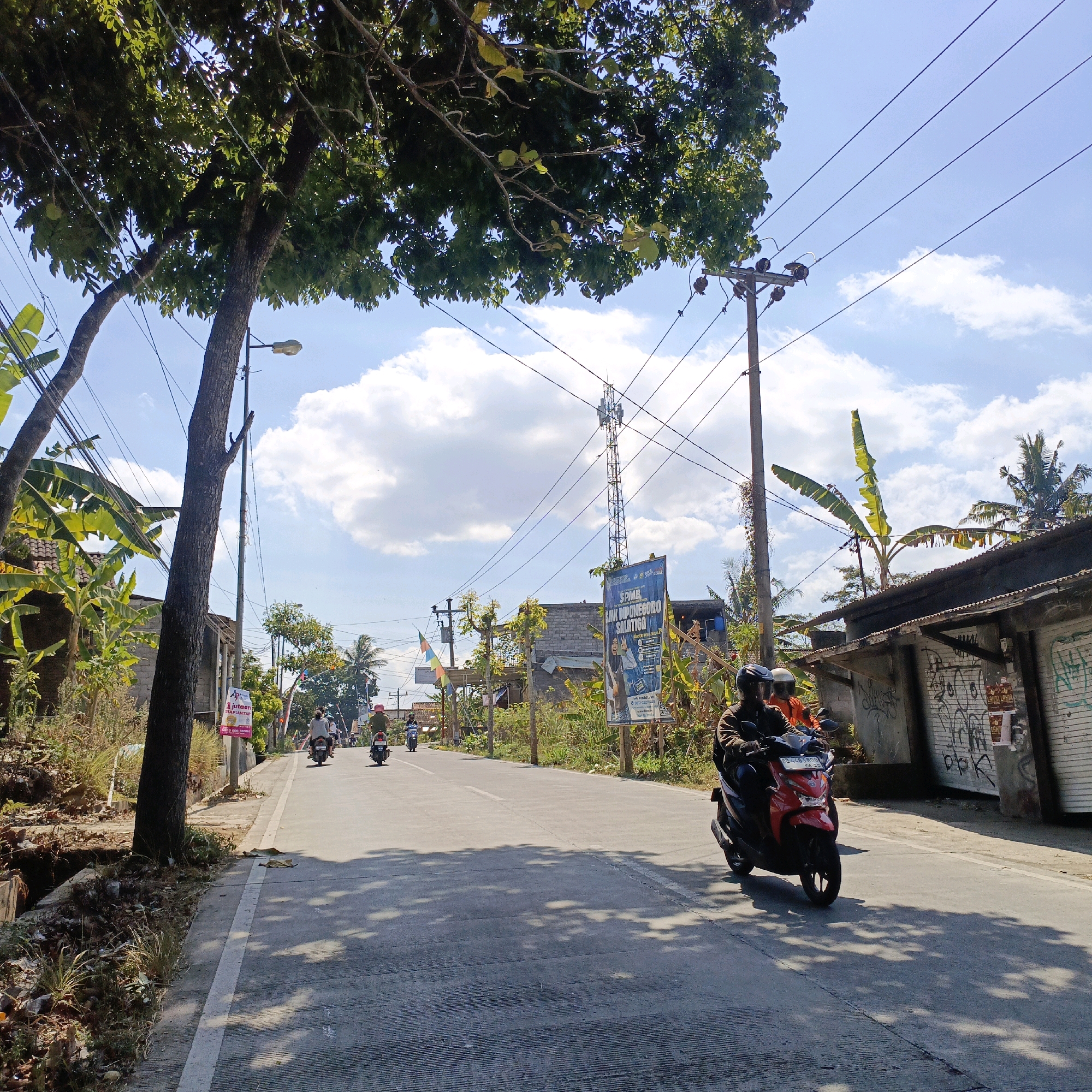This is a low-angle, medium-shot outdoor photograph taken on a sunny day in Salatiga, Indonesia. The scene depicts a paved road with a single lane of traffic heading away from the viewer. On the right side of the road, there is a row of buildings with corrugated metal roofs, some of which are covered in graffiti. A tall cell tower is visible in the distance behind these buildings. The left side of the road features a steep embankment with overgrown vegetation and a drainage ditch. Large trees with dark green foliage hang over the road on the left, casting dappled shadows on the pavement. Several power poles with overhead wires are present throughout the scene.
In the foreground, a red scooter is being ridden by two individuals wearing helmets. The driver is wearing a brown jacket, and the passenger is wearing an orange jacket. The scooter's license plate is visible. Further down the road, several other individuals are riding motorcycles, also with helmets. In the background, there are more buildings, trees, and a telecommunications tower. The sky is a clear blue with scattered white clouds. The overall impression is of a rural or semi-urban street scene in a tropical climate.
