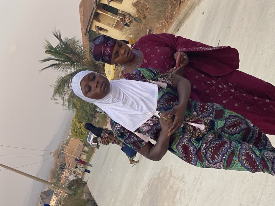 Two women are standing close to each other outdoors. The woman in the foreground wears a white headscarf and a patterned dress. The woman behind her has a patterned head covering and a maroon outfit. Both women have rings on their fingers. In the background, there are buildings, trees, and a few people further away. The sky is overcast.