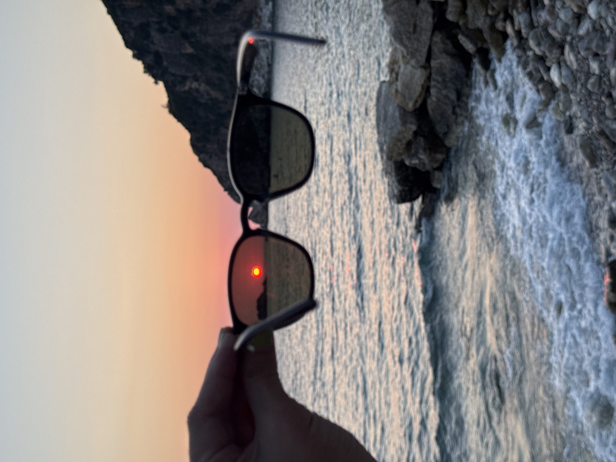 In the city of Perazića Do, Montenegro, a person's hand holds a pair of dark-rimmed sunglasses in the foreground. The lenses of the sunglasses reflect a vibrant sunset with a bright red sun positioned over the calm sea. The background reveals a dark, rugged hillside with vegetation, meeting the water. The sea has small waves breaking on a rocky shore, and the sky transitions from a light blue at the top to shades of orange and pink near the horizon.