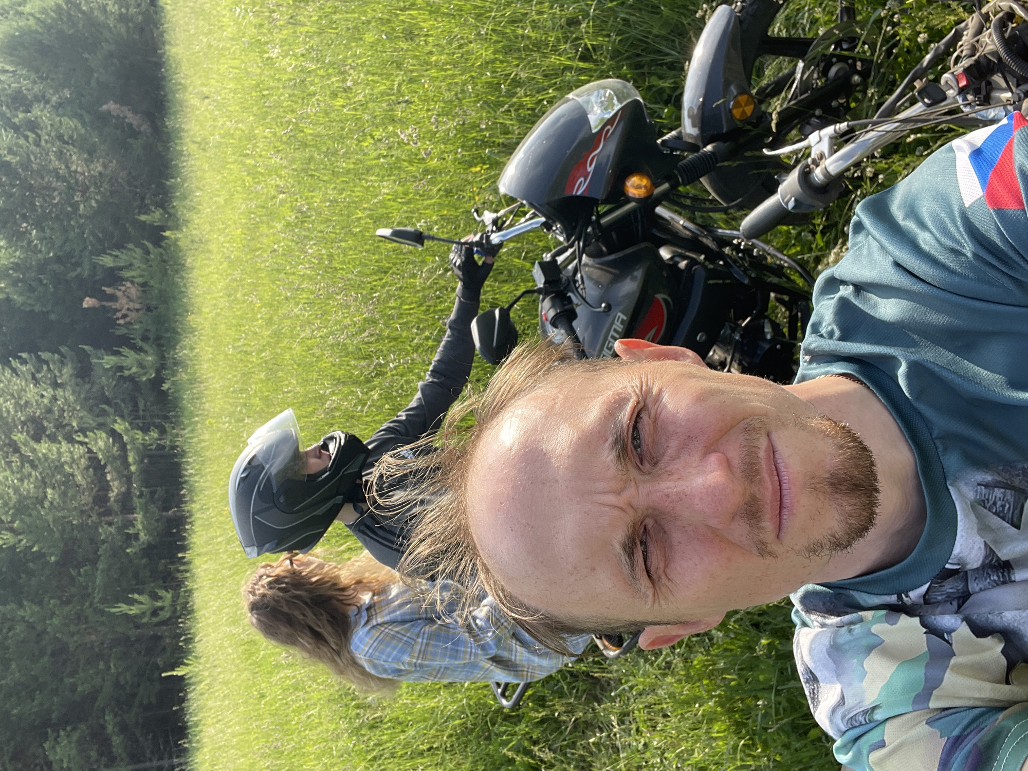 In the village of Nizhnii Bugrysh, Russia, a group of individuals is captured in a selfie-style photograph. In the foreground, a man with short, light brown hair and a goatee stares at the camera with a concerned expression. He wears a camouflage t-shirt. Behind him, a motorcycle is partially visible, with a rider wearing a black helmet and a dark jacket and a passenger wearing a blue plaid shirt. The motorcycle appears to be parked in a field of tall green grass, with a forest in the background. The overall scene suggests an outdoor excursion.