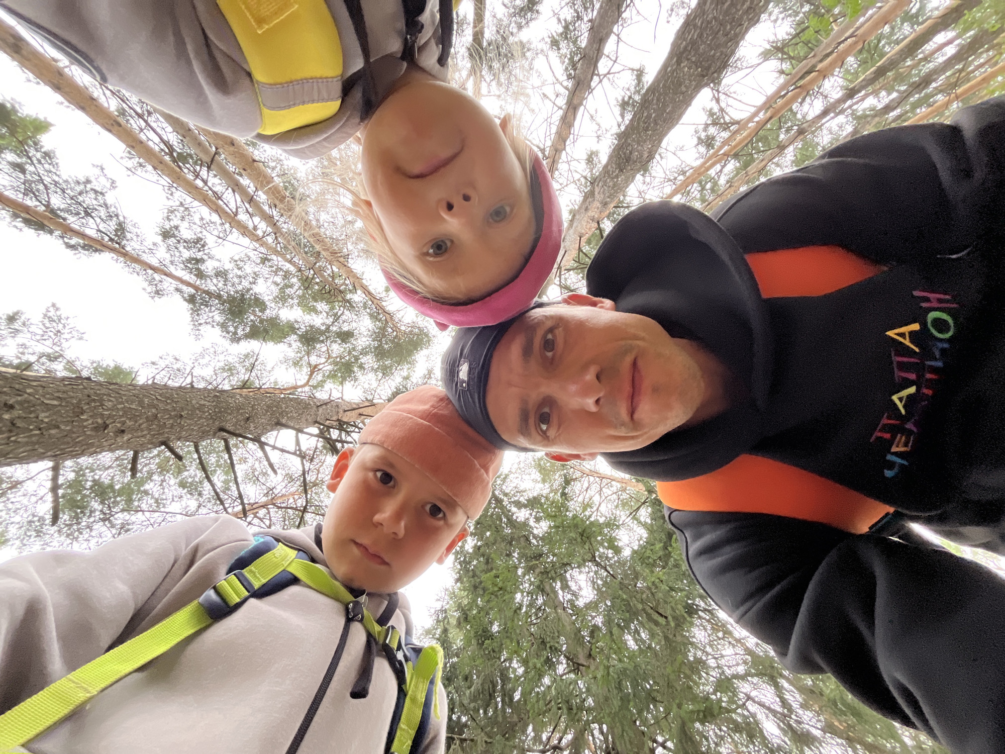A selfie taken from a low angle looking up at three people in a forest setting. The people appear to be a man and two children. The man is wearing a black hoodie with colorful lettering and an orange insert. One child is wearing a pink hat and a gray hoodie with a yellow safety vest. The other child is wearing a light-colored hoodie with a bright green safety harness and a peach-colored hat. The background consists of tall pine trees and a cloudy sky. The photograph was taken in the city of Chaikovsky, Russia.