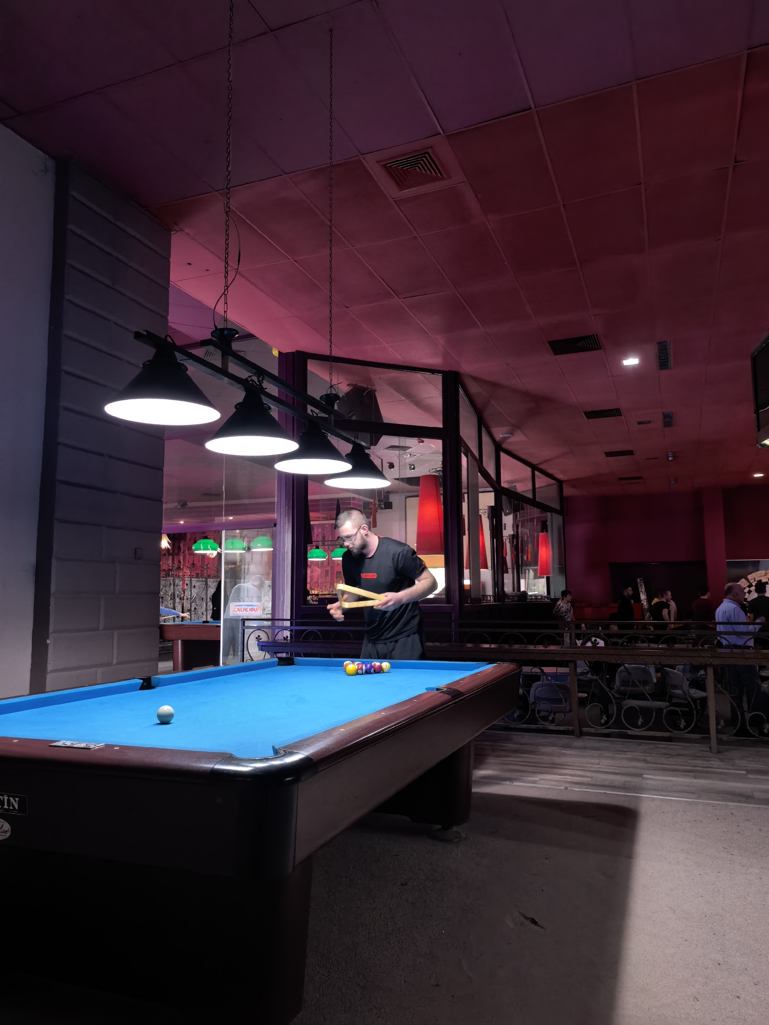 A photograph depicts the interior of a billiard hall in Plovdiv, Bulgaria. A pool table dominates the foreground, with a light blue felt surface and a set of billiard balls racked at one end. A man in a black t-shirt stands beside the table, holding a billiard bridge. Behind him, a glass partition separates the billiards area from a lounge-like area with tables, chairs, and other people. The lighting is dim, with a pinkish-red hue emanating from the ceiling and walls in the background. Above the pool table, a set of pendant lights illuminate the scene. The floor is light gray. Other billiard tables can be seen through the glass wall in the background. A bar can also be seen past the pool table.