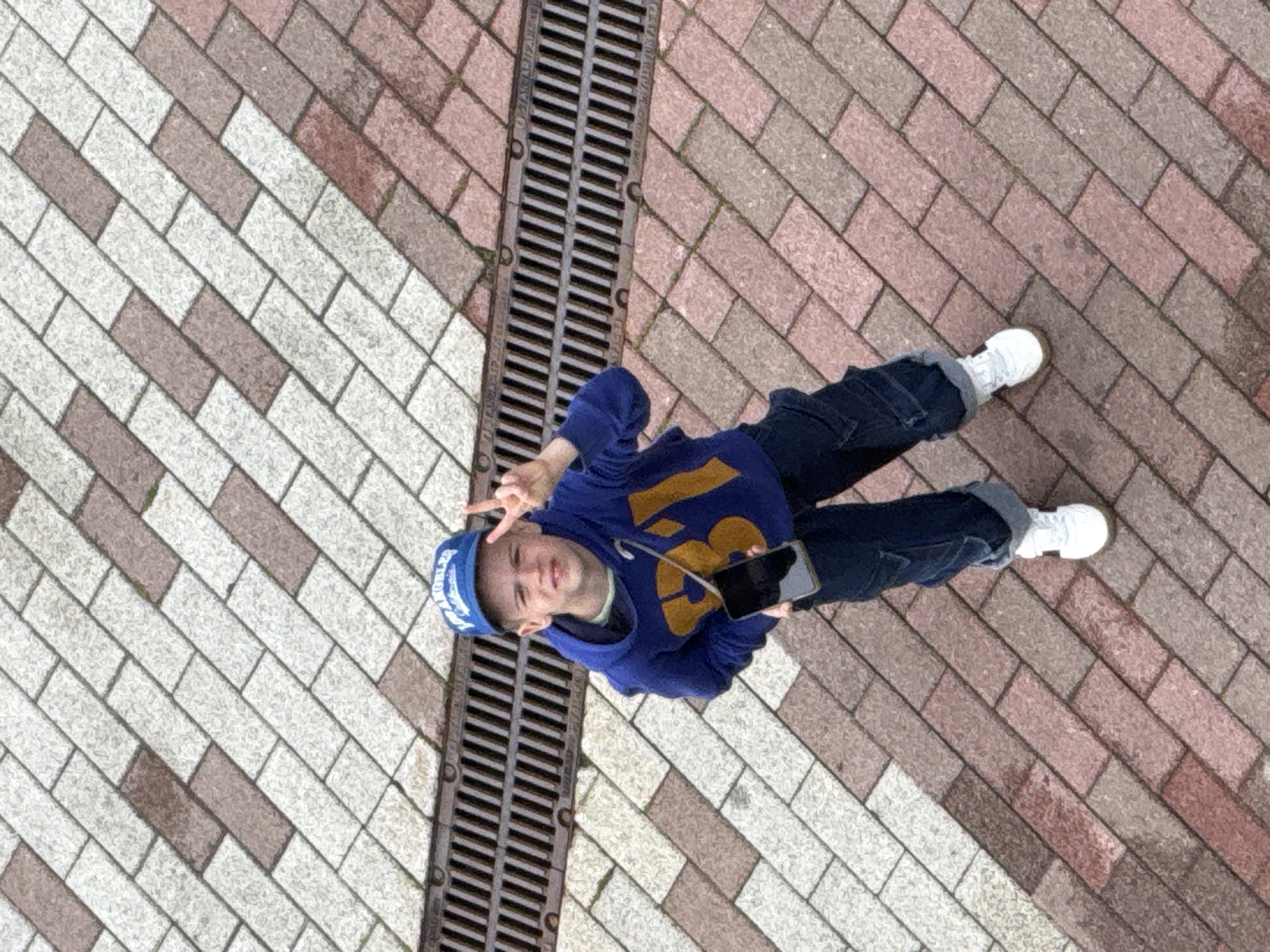 An outdoor, high-angle, full-shot photograph depicts a young Caucasian boy standing on a patterned sidewalk composed of beige and reddish-brown rectangular bricks. A dark metal grate runs horizontally across the mid-ground. The boy, with short, light brown hair, wears a blue baseball cap, a blue hooded sweatshirt with a large yellow "31" graphic on the chest, dark denim cargo pants, and white sneakers. He holds a black smartphone in his left hand and makes a peace sign with his right hand, looking towards the camera with a slight smile. The lighting is overcast, suggesting a cloudy day. The specific location in Moscow, Russia, cannot be determined from the visual information alone.