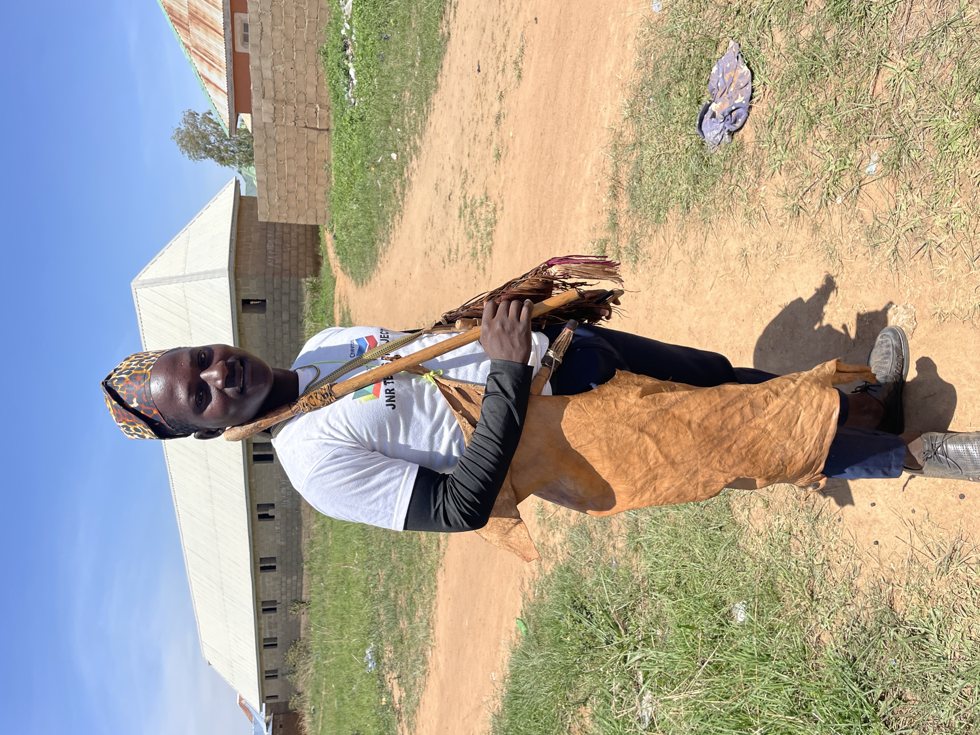 A man stands on a dirt path in Durumi, Nigeria. He wears a white t-shirt with black sleeves, blue pants, and dark shoes. On his head is a patterned scarf. He has a brown leather hide slung across his torso, and carries sticks and dried plants in his hands. In the background are block buildings with corrugated metal roofs and patches of grass. The sky is clear blue.
