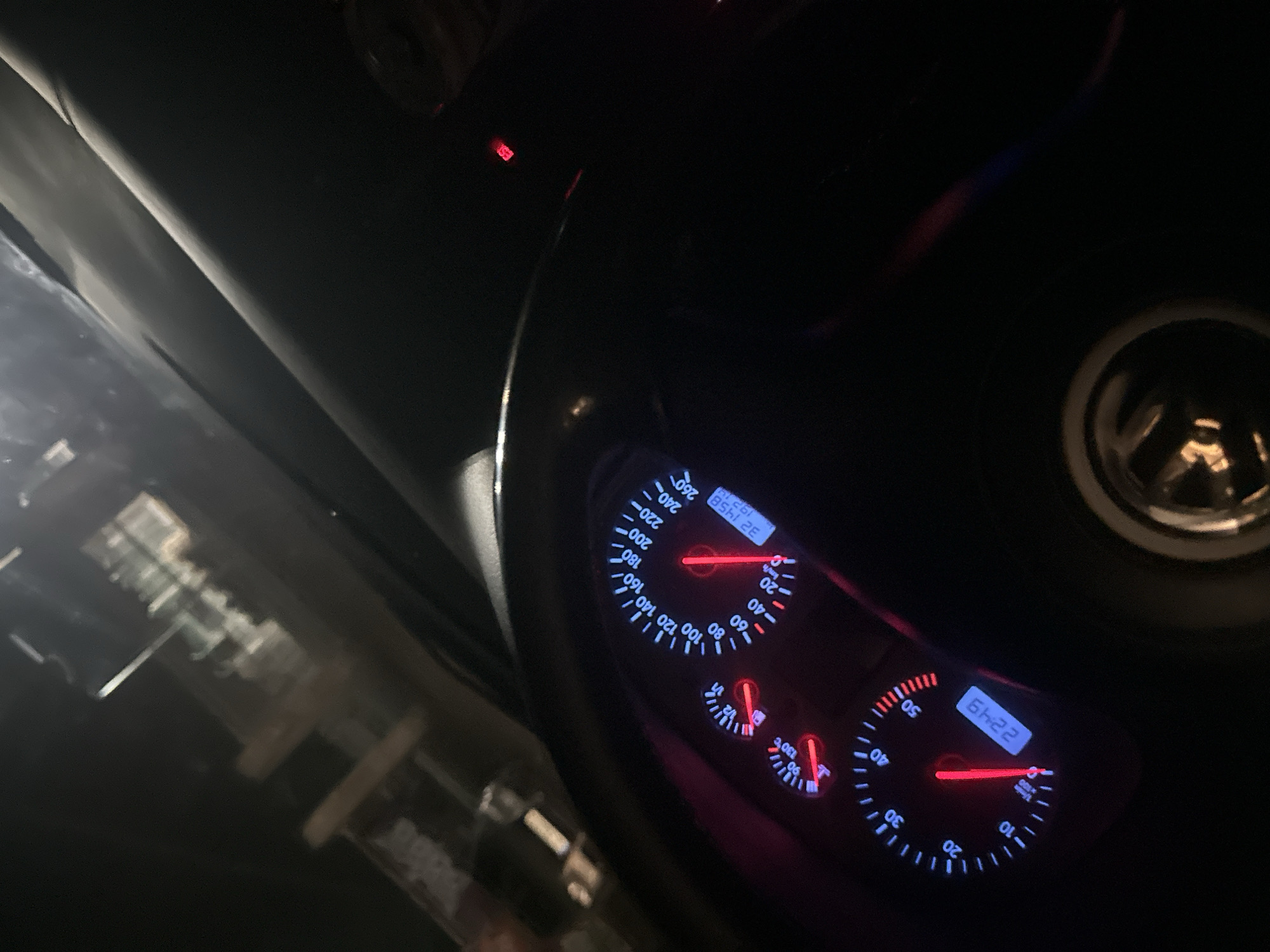 The image shows a car's dashboard at night. The speedometer indicates approximately 180 km/h, and the tachometer shows the engine speed is around 1,000 RPM. A digital display reads "22:49" and the odometer displays "32 1458" and "19214". The steering wheel is visible, and the vehicle appears to be in motion. The exterior environment is dark, with blurred lights suggesting an urban area at night. This scene is unlikely to be in the city of Karnobat, Bulgaria, as it would be unusual to be going this fast within city limits.