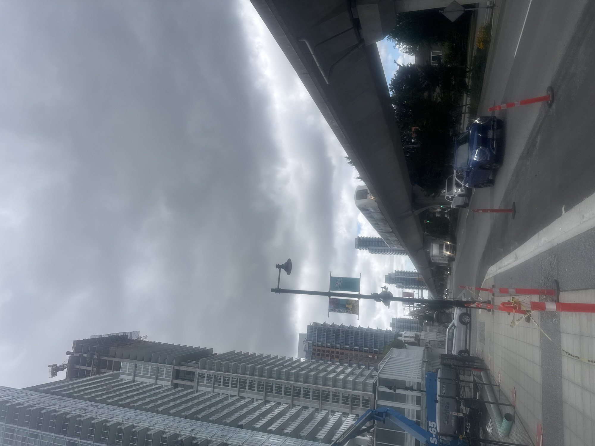 This is a medium shot, eye-level, daytime, outdoor, vertical, full-color, slightly blurry image taken from a sidewalk in Coquitlam, Canada, looking towards a multi-lane road with a SkyTrain line overhead. The sky is heavily overcast with dark gray clouds. On the left side of the frame, a tall, unfinished skyscraper dominates the scene, with construction equipment and materials visible at its upper levels. Below the skyscraper, a blue construction lift is parked. The sidewalk in the foreground is paved with gray tiles and has red and white construction cones with yellow caution tape.
The road in the mid-ground and background has several vehicles, including a dark blue SUV in the right lane, a white SUV and a white pickup truck further ahead. A white freight truck is also visible further down the road on the right. The SkyTrain line, an elevated concrete structure, runs diagonally across the upper right portion of the image. A SkyTrain car is visible on the tracks, approaching from the left. Trees and some residential buildings are visible on the right side of the road, beyond the SkyTrain line. Two lampposts with banners are positioned along the sidewalk. The overall impression is one of urban development and ongoing construction under a gloomy sky.
