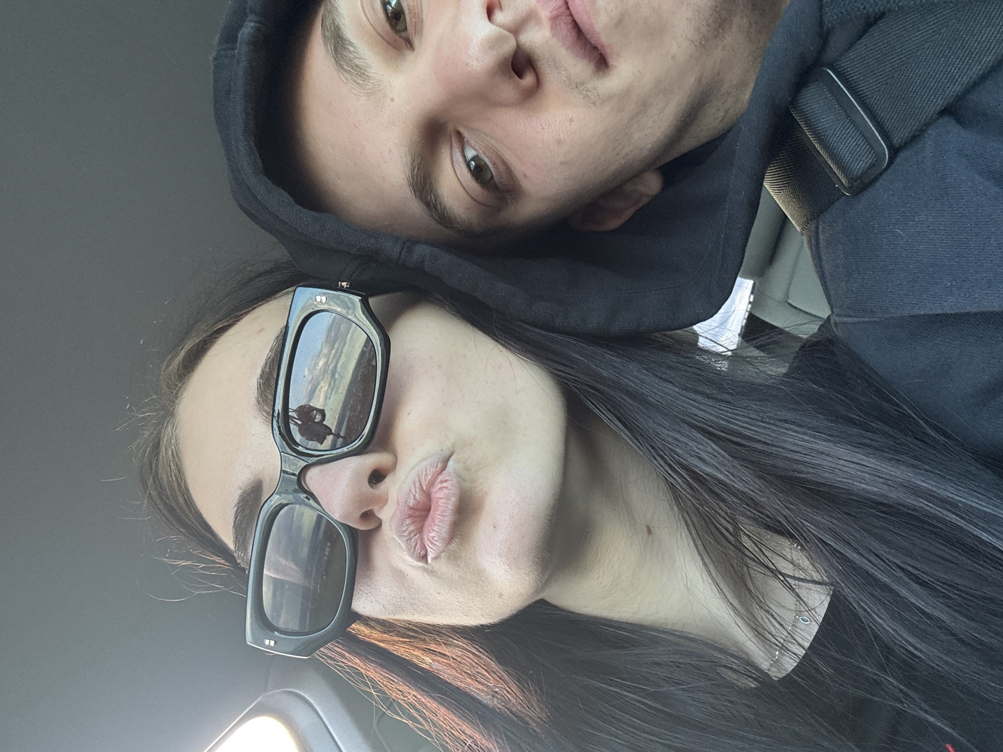 A close-up selfie shot indoors, presumably inside a car, shows two individuals. A young woman with long, straight, dark hair is positioned at the bottom of the frame, wearing black, oversized sunglasses and pursing her lips. Above her, a young man with short hair is wearing a black hooded sweatshirt. The angle suggests the woman is holding the camera, and the man is leaning towards her. Reflections visible in her sunglasses suggest an outdoor environment. Given the provided location of Burgas, Bulgaria, the reflection likely shows elements of the city or its surroundings.