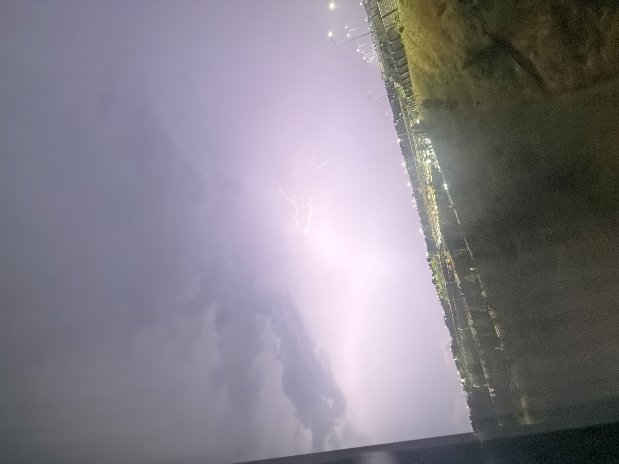 The image captures a dramatic lightning strike illuminating the sky over a cityscape, likely at dusk or night. The lightning forks across the purplish-grey sky, creating a bright, almost ethereal glow. Below, the city lights are visible, suggesting a built-up area. A dark foreground element, possibly a window frame or barrier, partially obstructs the view on the left side of the image. The rest of the scene shows a landscape with some greenery and streetlights lining what appears to be a road or an elevated infrastructure, with a fence or barrier running along the side. The overall impression is one of a powerful natural phenomenon occurring over a populated area.