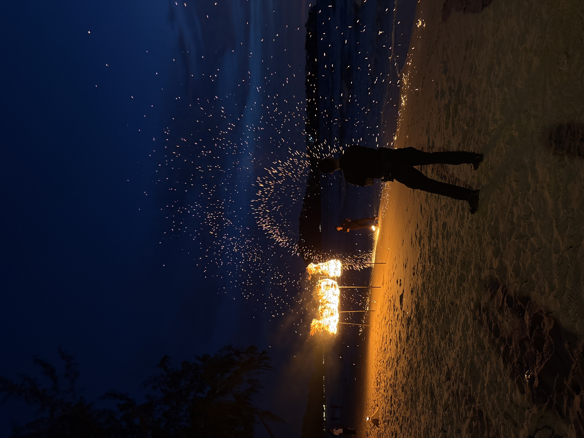 The image depicts a night scene on a sandy beach in Choeng Thale, Thailand, likely during a performance or event. In the foreground, a person stands with their back to the camera, silhouetted against the scene. They are wearing dark clothing and dark shoes. To the left of the frame, several structures are engulfed in bright orange flames, illuminating the sand and creating a warm glow. These flame structures are supported by thin poles. Behind these flames, sparks are being ejected upwards and outwards, creating a showering effect that arcs across the dark blue night sky. The ocean is visible in the background, with a faint outline of land on the horizon. The sky is a deep indigo, with scattered clouds. The overall impression is one of a dynamic and visually striking performance occurring on a beach at night.