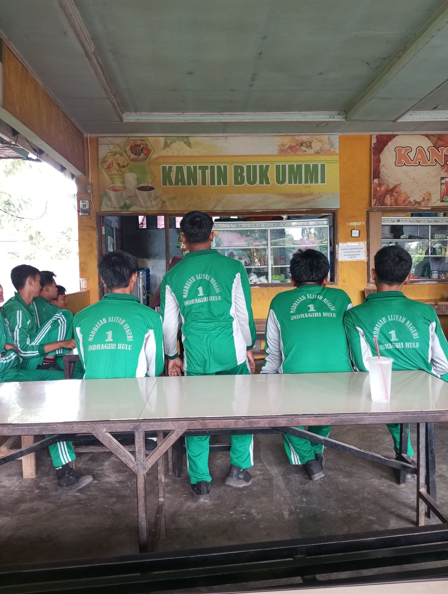 A group of young males, likely students wearing matching green tracksuits with white trim and the inscription "MADRASAH ALIYAH NEGERI 1 INDRAGIRI HULU" on the back, are seated at a metal table in what appears to be an outdoor canteen or food stall. The canteen has a yellow and orange mural on the wall depicting food items and the text "KANTIN BUK UMMI." Some of the students are facing away from the camera, while others are partially visible from the side. They are seated on benches or chairs. A white plastic cup with a red straw is on the table in front of two of the individuals. The floor is concrete, and the environment suggests a casual, open-air setting, consistent with a canteen in Pematang Reba, Indonesia. The lighting suggests it is daytime, with natural light filtering in from the left.