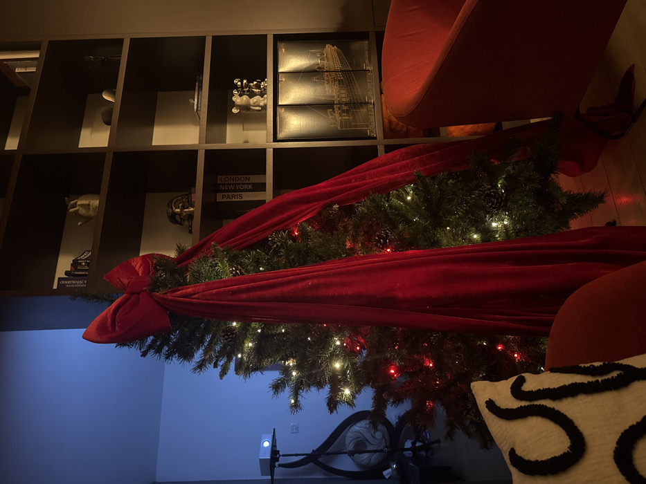 An indoor scene depicts a modern living area decorated for the Christmas season, observed at a slightly elevated angle. The specific location within the city of Warsaw is not discernible from the image.
The central focus is a tall, artificial Christmas tree adorned with numerous small white and red string lights. A large, crimson velvet bow is positioned at the top of the tree, with wide, matching velvet ribbons draped vertically down its sides, concealing parts of the tree. Several pinecones are visible among the green artificial branches.
To the right of the Christmas tree, a dark-colored, multi-tiered bookshelf unit is against a dark wall. From top to bottom, visible items on the shelves include:
* An off-white, stylized human head sculpture and other small, indistinct objects on the top shelf.
* A golden or bronze ornate mask, possibly a Venetian or theatrical style, on the second shelf.
* Three books with visible spines labeled "LONDON," "NEW YORK," and "PARIS" on the third shelf.
* Two light-colored, stylized bear-like figures (resembling KAWS figurines) on the fourth shelf.
* Three large, dark-toned boxes or books featuring a gold-outlined illustration of a ship (identifiable as the Titanic) on the bottom shelf.
On the far left, a white digital projector rests on a slender, black-legged stand. The wall behind the projector is illuminated by a cool blue ambient light, contrasting with the warmer, yellowish light in the rest of the room.
In the foreground, the upper portion of a red upholstered armchair is visible on the right. On the far left, a partial view of another red upholstered seating element is present, topped with a white cushion featuring black, abstract, wavy patterns. The floor beneath the tree and seating is a light-toned wooden surface. No individuals are present in the scene, and no specific actions or interactions are depicted. The overall environment suggests a contemporary residential or hospitality interior.