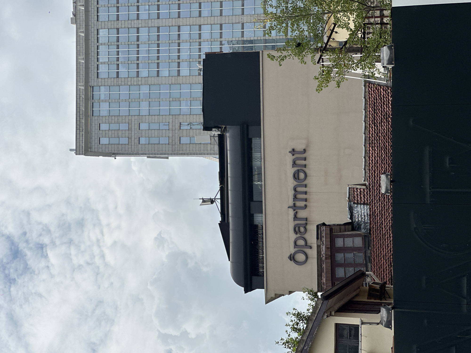 The image shows a building with the word "Ôpartment" in large, dark lettering. It is a multi-story building with a beige facade. Above the "Ôpartment" sign, there is a dark, curved structure, possibly a roof or an architectural element. In the background, a taller, modern building with many windows is visible, suggesting an urban setting. To the left, there is a smaller, older building with a tiled roof and wooden window frames. Trees are also present, with some foliage visible on the right side of the image. The sky is overcast with clouds. The scene appears to be a street view in a city. This building is located in Shanghai, China.