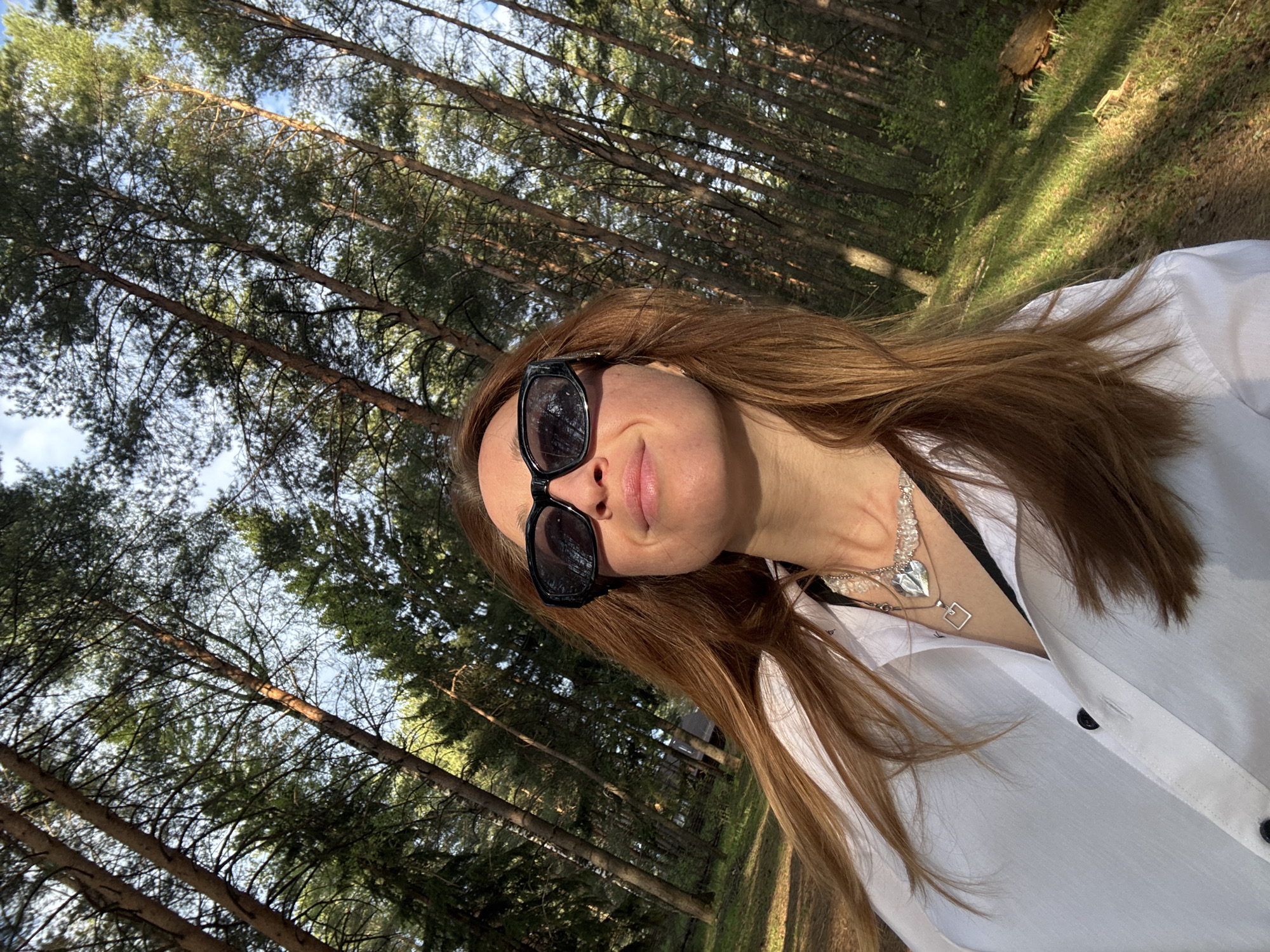 A selfie depicts a woman wearing sunglasses and a white button-down shirt, with a silver necklace visible. She is positioned against a backdrop of tall trees, likely in a forested area. The location is Прикамский, Russia. Sunlight filters through the trees, creating varied light and shadow patterns. The image appears tilted, with the trees leaning towards the left.
