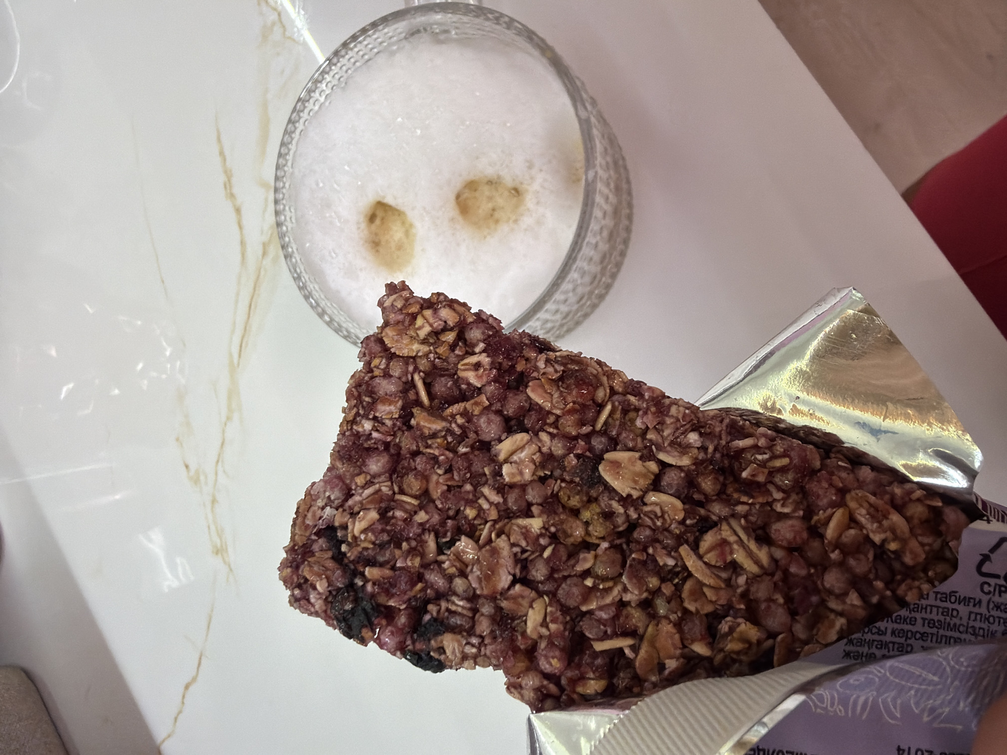 The image displays a close-up, overhead view of a textured granola or cereal bar and a glass of foamy beverage on a white, marble-patterned surface. The bar, a dark reddish-brown, is partially unwrapped from its silver and purple foil packaging, revealing a dense mixture of grains, dried fruit, and potentially nuts.
Next to the bar is a clear, ridged glass containing a light, frothy drink, likely coffee or a latte, with two darker spots on the foam resembling eyes. The white surface features subtle golden-brown veining, characteristic of marble or a similar material. In the background to the right, a small portion of a red object, possibly furniture, is visible.
The scene suggests an individual is enjoying a snack or light meal during the daytime in an indoor setting, consistent with the provided context of Sochi, Russia. Visible text on the bar's packaging, in Cyrillic, includes words like "natural," "sugars," "gluten," and "nuts," along with the number "2014," likely a date related to manufacturing or expiry.