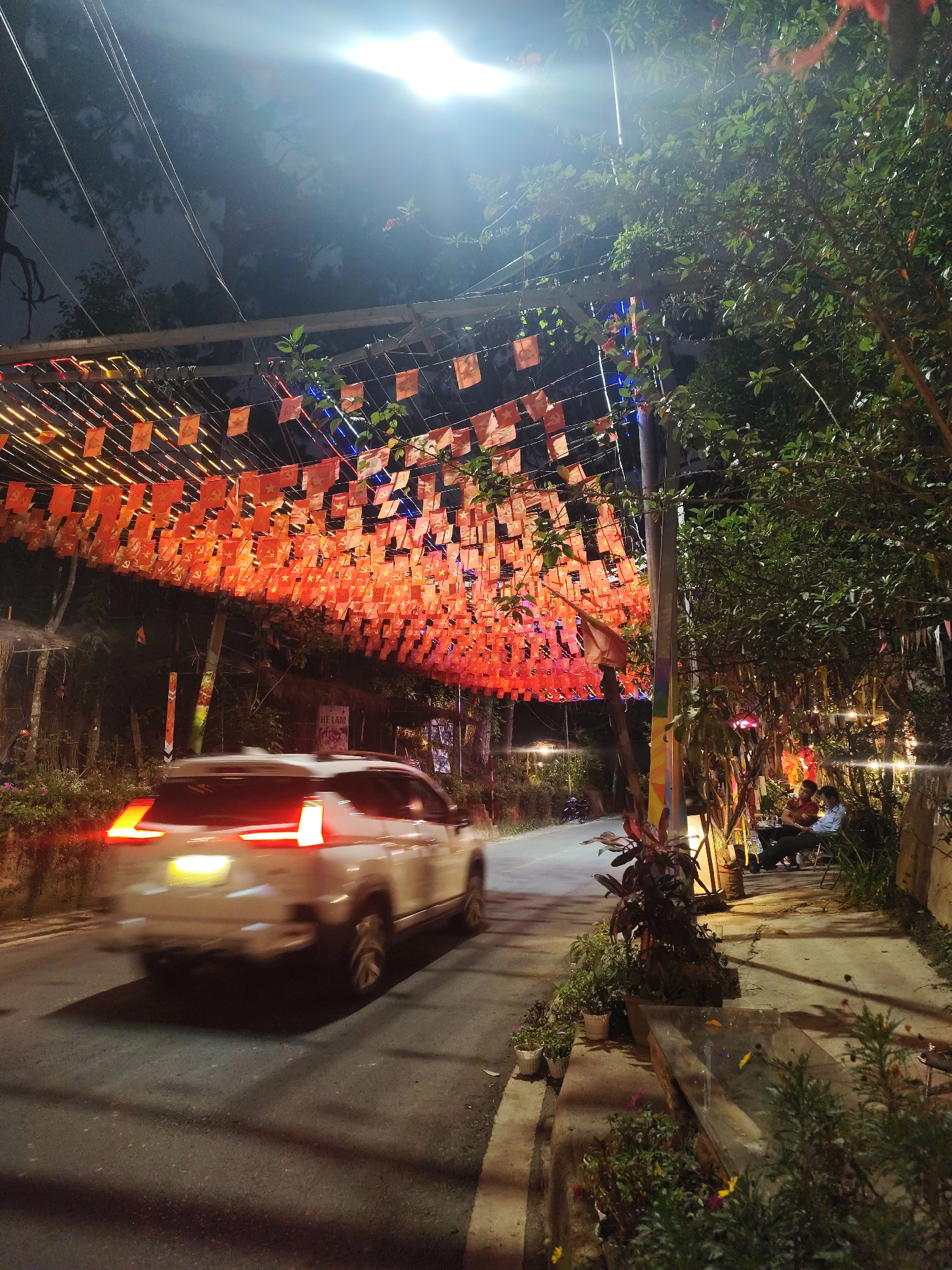 The photo shows a street scene at night in Da Lat, Vietnam. A white SUV moves along the asphalt road, causing motion blur. Above the road, a rectangular metal frame stretches across the street. Numerous small, orange flags hang from the frame, creating a canopy effect. A street light illuminates the scene from above.
To the right of the road, a sidewalk is present. People are seated at tables, suggesting an outdoor café or dining area. Lush vegetation and trees line the edge of the sidewalk. The background is dark, indicating night or low lighting.