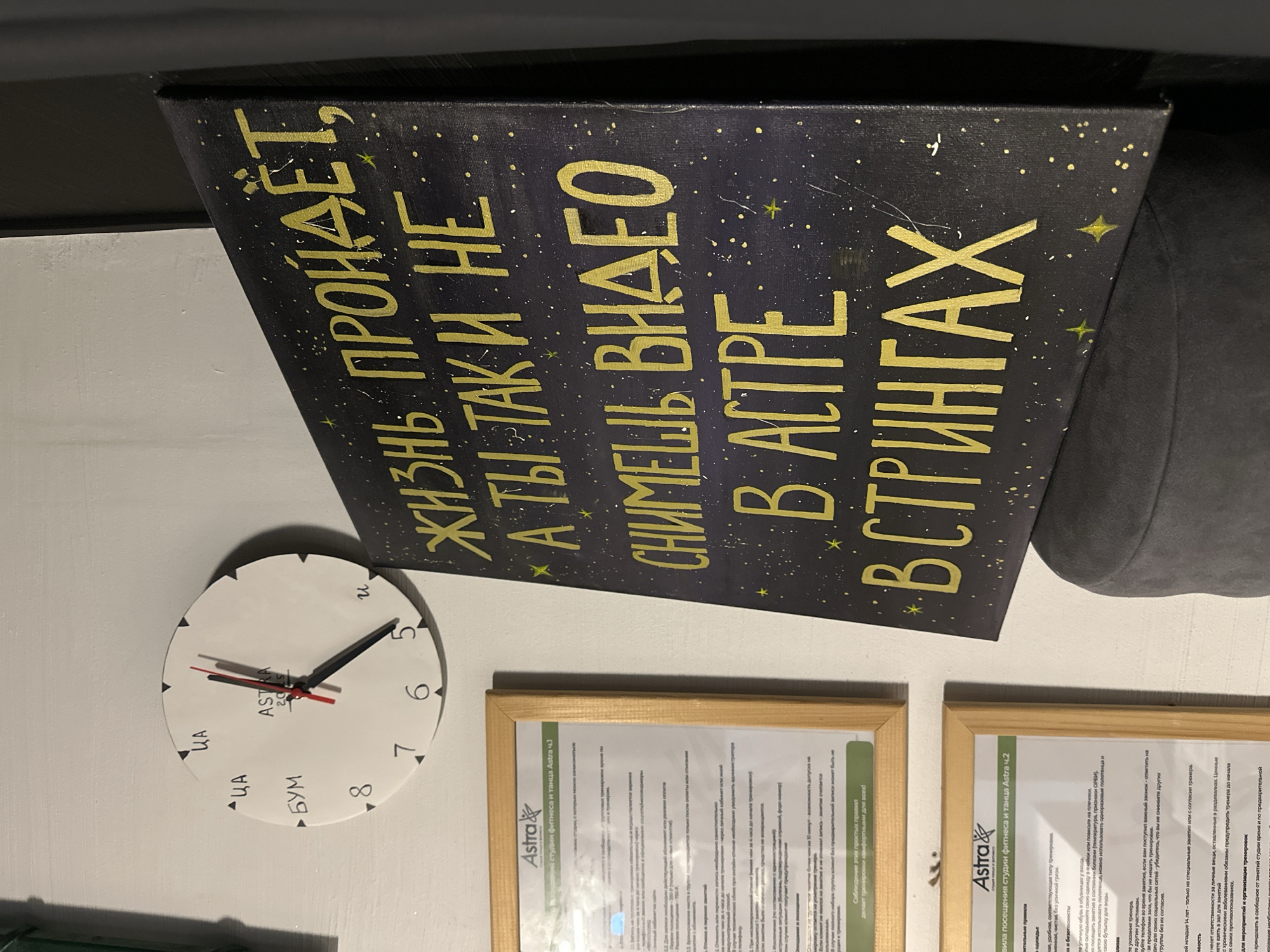 This media file is a photograph taken in Vladimir, Russia, likely indoors. The main subjects are a wall clock and a black canvas with golden lettering. The canvas is tilted, displaying a message in Russian. The clock is white with black markings and hands, showing approximately 5:30. The wall appears to be painted white.
The canvas has a starry background and the golden text, when read from bottom to top and left to right, appears to be a motivational or humorous quote. The visible parts of the text include phrases like "ЖИЗНЬ ПРОЙДЁТ," "А ТЫ ТАК И НЕ СНИМЕШЬ ВИДЕО," and "В АСТРЕ В СТРИНГАХ".
There are also two framed documents on the wall below the canvas. These documents have the "Astra" logo and appear to be rules or guidelines for a fitness and dance studio. One document is titled "Правила посещения студии фитнеса и танца Astra 4.1" and the other "Правила посещения студии фитнеса и танца Astra 4.2".
The overall impression is of an interior space, possibly a studio or a waiting area, decorated with artistic elements and informational materials. There are no people visible in the image. The lighting suggests it could be daytime, but the exact time of day is not clear from the illumination alone. The scene is static and does not capture any ongoing action.