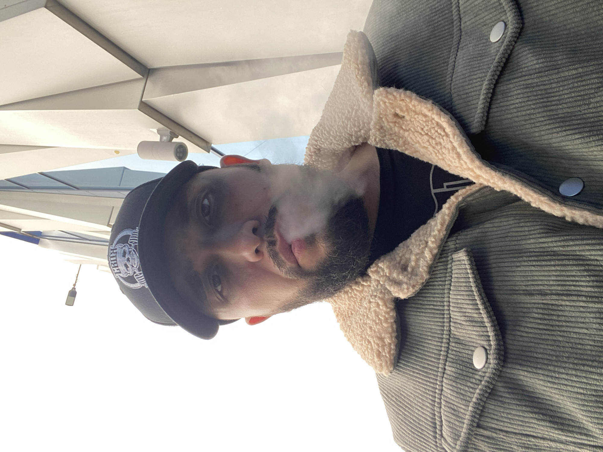 A man with a beard and a black baseball cap with a white logo is exhaling smoke. He is wearing a green corduroy jacket with a beige shearling collar. The photo is taken from a low angle, looking up at the man. In the background, there is a building with a white, geometric ceiling. There is a surveillance camera attached to the ceiling. A pole and a flag of white and blue are visible on the left. It is not possible to identify the location as Chaikovsky, Russia, based on the provided image.