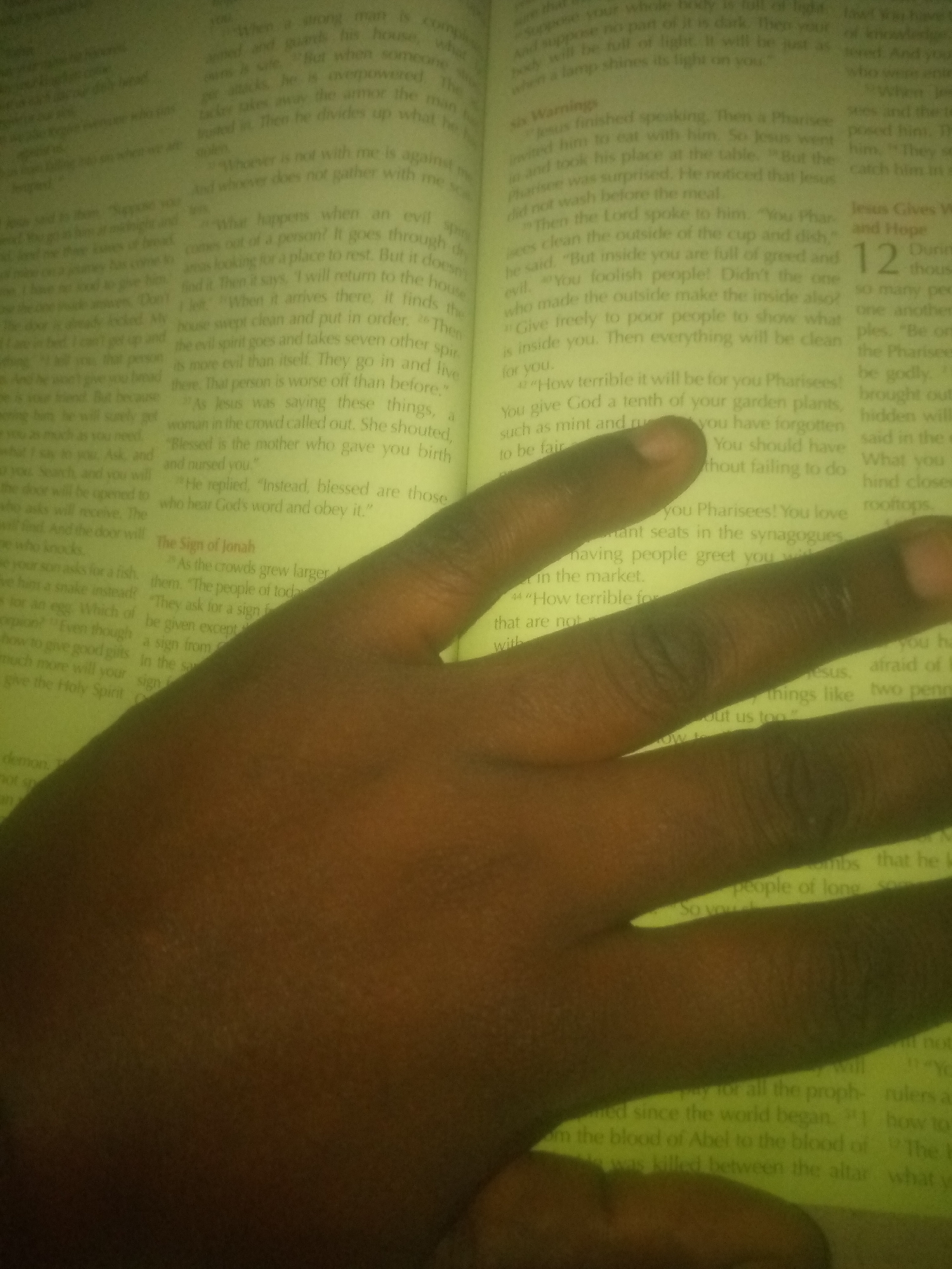 The image shows a close-up of a person's hand resting on an open book, likely a Bible, with text visible. The hand is positioned in the lower right portion of the frame, obscuring some of the text. The book's pages are illuminated by a warm, yellow light, suggesting ambient lighting conditions. The text in the book is in English and appears to be a religious or biblical passage, with some phrases like "When a strong man," "what happens when an evil spirit," and "How terrible it will be for you Pharisees" being partially legible. The image is slightly blurry, particularly the text in the background. There is no discernible information within the image to suggest a location in Jauro Yinu, Nigeria, or any other specific geographical setting. The scene is static and focused on the interaction between the hand and the book.