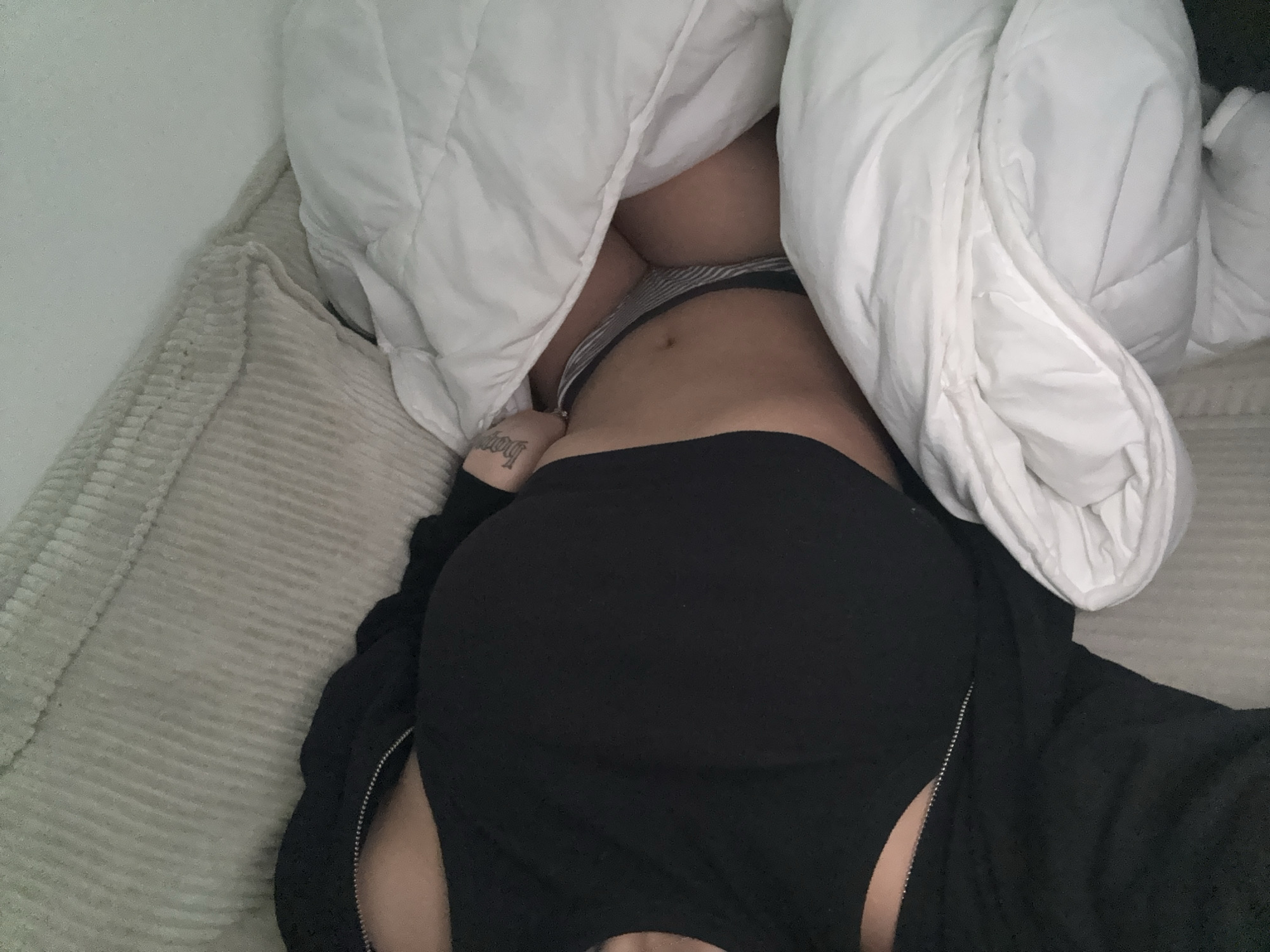 A person lies on a beige corduroy couch with a white comforter pulled up over their body. The individual wears black bottoms and a black zippered jacket. A tattoo is visible on the person's left side. The image appears to be taken indoors. I cannot determine the location in Mönchengladbach, Germany, from the image.