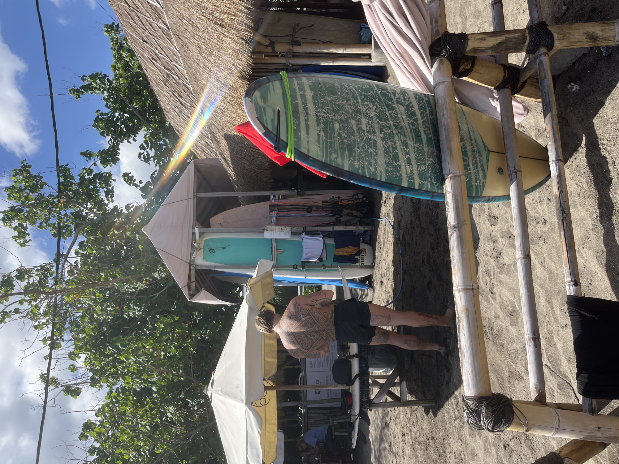 The image depicts a coastal scene in North Kuta, Indonesia. Several surfboards are stacked in a rack near a structure with a thatched roof. A man with a large tattoo on his back stands near the surfboard rental business. A white umbrella is present over the establishment. Patrons sit beneath the umbrellas. The ground is sandy, and trees are visible in the background, with a blue sky and some clouds overhead.