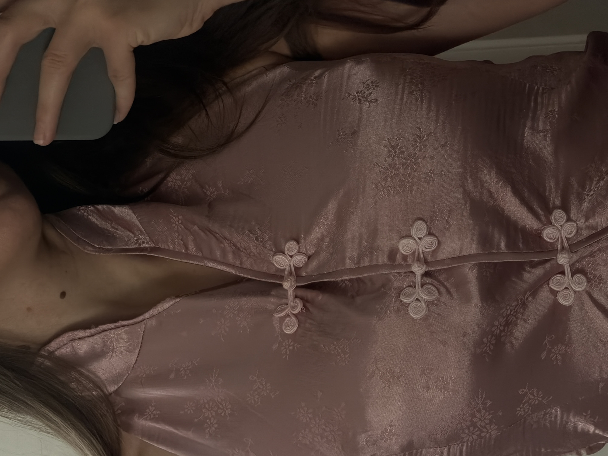 The image shows a close-up of a person wearing a pink satin halter top with a Mandarin collar and decorative knot buttons. The fabric has a subtle floral pattern. The person's neck, shoulder, and upper chest are visible. The person is holding a grey mobile phone in their right hand, partially obscuring their face and hair. The background is indistinct. There is no indication of the city of Sharm El Sheikh, Egypt, in the image.