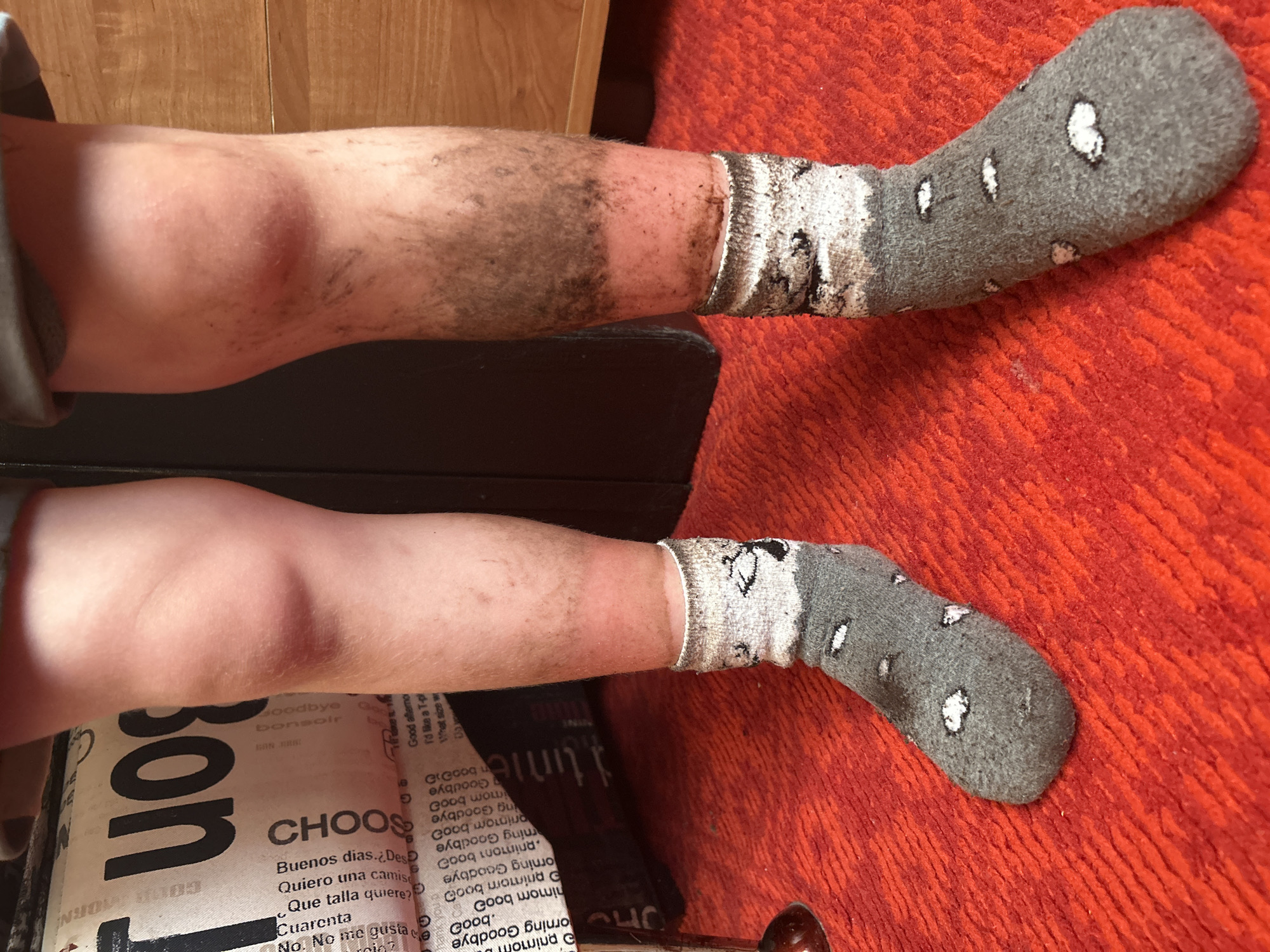 The image shows two legs resting on an object with text. The upper leg is dirty and wearing a gray sock with white spots. The lower leg is less dirty and also wearing a gray sock with white spots and cartoon images. The legs are resting on an object printed with text like "Goodbye, bonsoir" and "CHOOS". The background features a wooden surface and a textured orange surface. There is no evidence to determine the location as Noviy, Russia.
