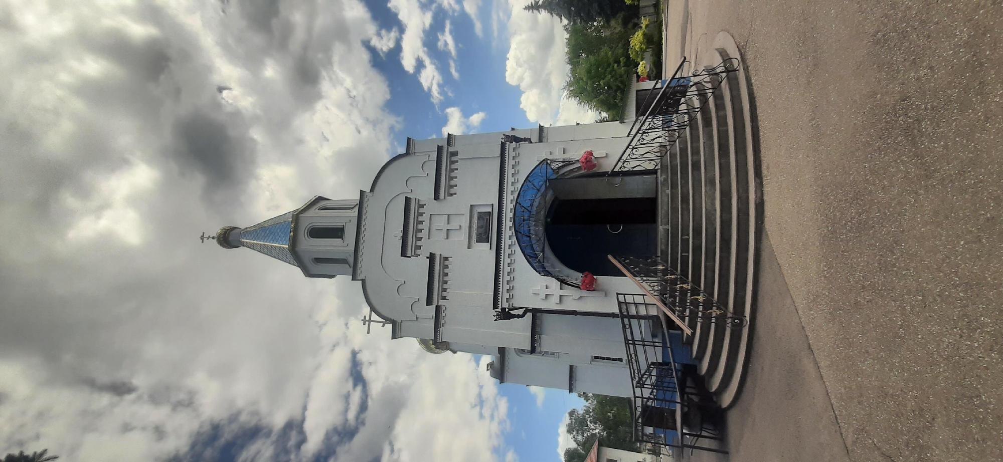 The image depicts the exterior of a white Orthodox church with a blue-tiled, pointed roof topped by a cross. The church features a prominent arched entryway with a blue awning and decorative red flowers on either side. A wide, curved staircase with black metal railings leads up to the entrance, with a handicap-accessible ramp also visible on the left. The church is situated in an area with a paved courtyard, surrounded by green trees and a cloudy sky. The location is identified as the city of Yessentukskaya, Russia. There are no people or animals visible in the image.