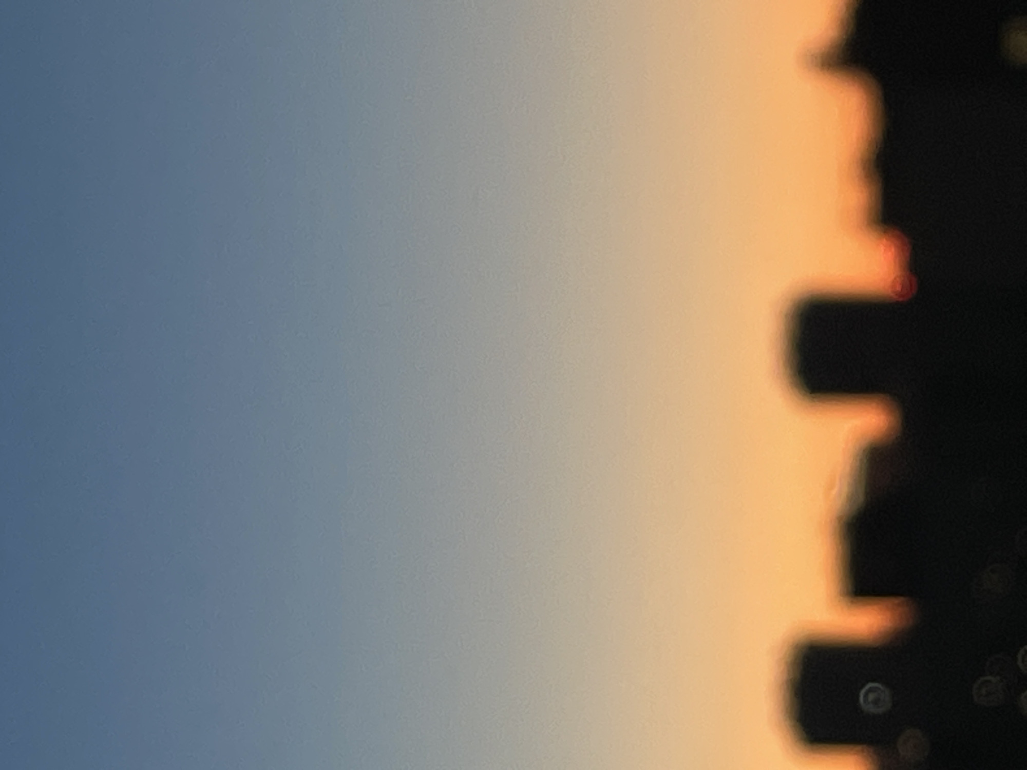 The image shows a blurred silhouette of buildings against a gradient sky transitioning from a pale blue at the top to a warm orange near the horizon. The buildings appear to be part of a cityscape. There are no people or discernible actions depicted. The scene is devoid of specific details due to the blurriness, making it impossible to ascertain the exact location within Sinyavino, Russia, or to identify any specific landmarks. The overall impression is that of a distant urban skyline at dusk or dawn.