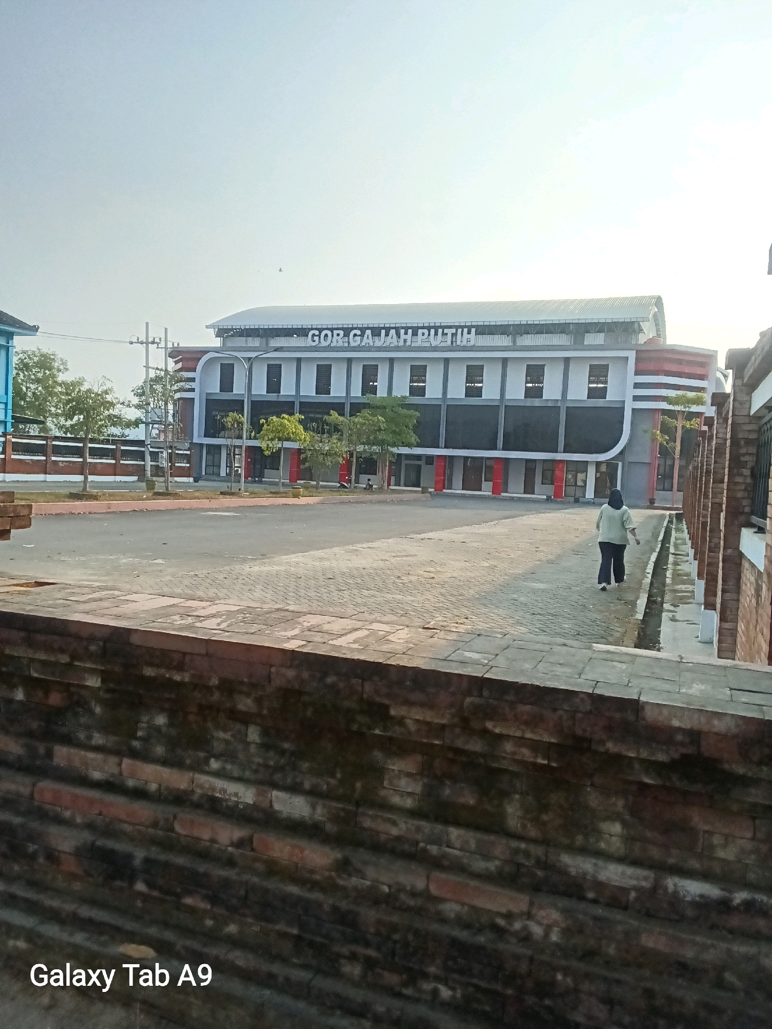 A wide-angle outdoor shot captures the "GOR GAJAH PUTIH" sports hall in Trenggalek, Indonesia. The building, a modern structure with a curved white roof and a prominent sign, features multiple windows and red accents. In front of the hall, a paved outdoor area is sparsely populated. A lone individual, seen from behind, walks away from the camera on the right side of the paved area, wearing a light-colored top and dark pants. The foreground is dominated by a textured, reddish-brown brick wall. The sky above is a pale, hazy blue, suggesting it is either early morning or late afternoon. The overall scene conveys a sense of public space that is quiet and relatively empty.