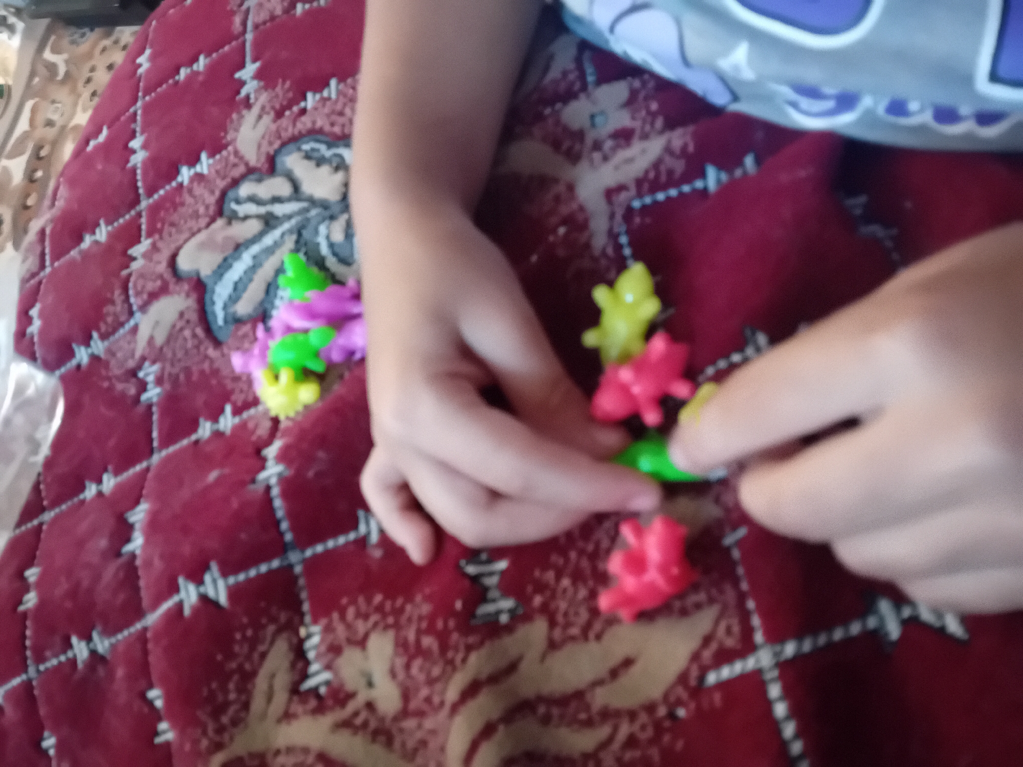 A child's hands are interacting with small, brightly colored toy animals on a red, patterned fabric surface, possibly a blanket or bedspread. The toys appear to be dinosaurs or similar creatures, in colors such as pink, green, and yellow. The fabric has a quilted pattern with a diamond grid and a floral or abstract motif. The background is blurred but suggests an indoor setting. There are no discernible landmarks or features that would indicate a specific location in Chuhuiv, Ukraine.