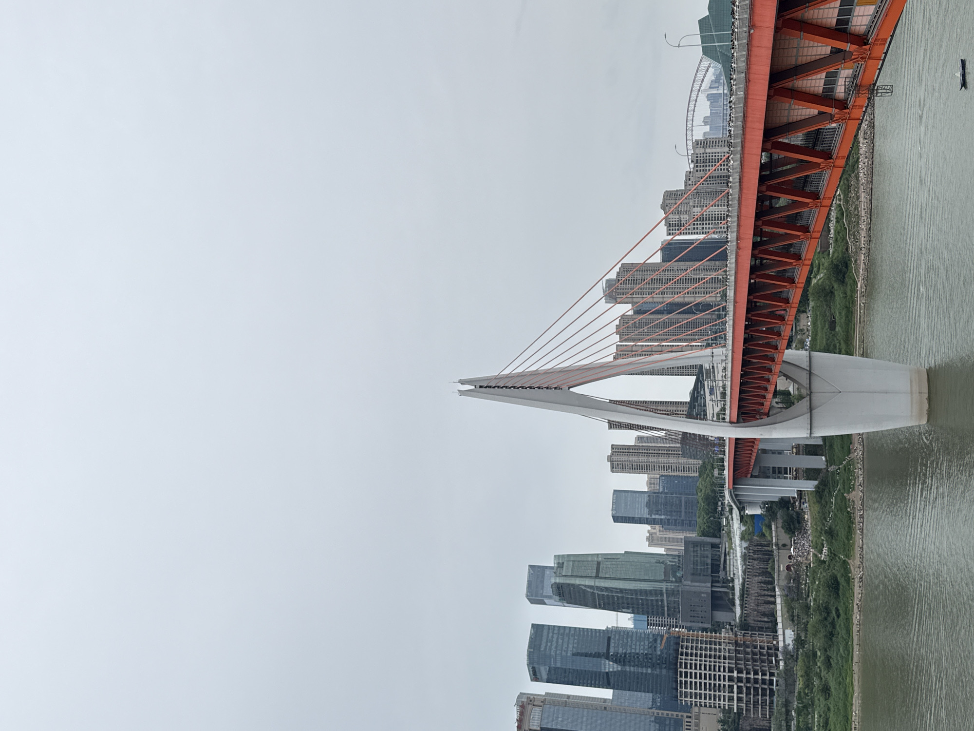 The image depicts the Dongshuimen Bridge, an architectural landmark in the Yuzhong District of Chongqing, China. The scene is a view of the city and bridge from a vantage point, likely along the Jialing River.
The bridge itself is a vibrant orange color with white supports, and is suspended over the water. Skyscrapers are visible on either side of the river. The sky is overcast, suggesting a grey or rainy day. The riverbank is lined with greenery and buildings. The bridge's architectural design is evident in its cables and structural components.
