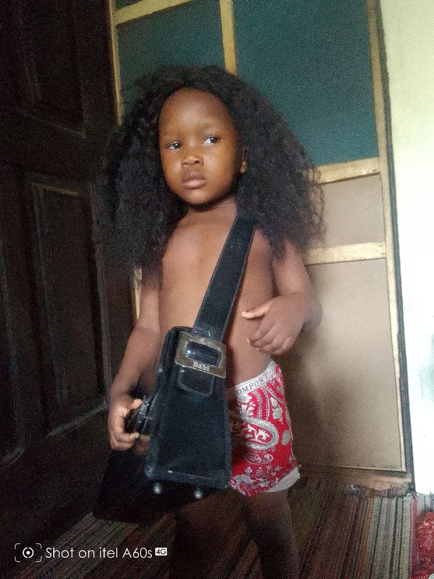 A young child stands indoors, partially clothed in red paisley boxer shorts, wearing a dark curly wig, and carrying a black bag slung over their shoulder. The bag's buckle reads "Rabi." The child is positioned near a dark doorway and a light-colored wall with dark trim. The photograph was taken using an itel A60s 4G phone. The location of the photo within Ikeja, Nigeria, is undetermined based on the image alone.