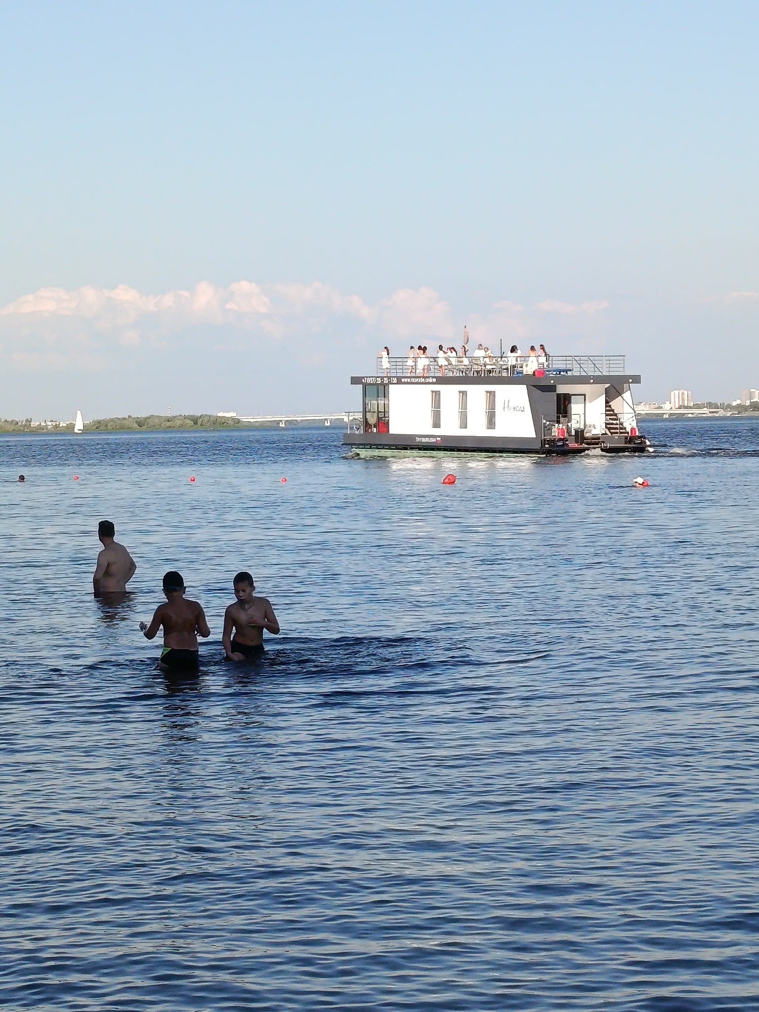 In a wide shot of a blue lake on a sunny day, three boys are wading in the water. One boy is closer to the camera and is facing away from it, looking at the other two boys. The other two boys are further back in the water and appear to be talking to each other.
In the background, a houseboat with people on its deck is moving across the water. The houseboat is white and black and has several windows. There is text on the side of the houseboat that reads "+7 (495) 25-25-135 www.mrade.online". The people on the deck are dressed in white clothing.
The sky is a clear blue with a few white clouds. The sun is shining, casting shadows on the water. The overall mood of the image is peaceful and relaxed, suggesting a summer day at the lake. There are a few buoys visible in the water, marking a swimming area. In the far distance, there is a bridge and city skyline.