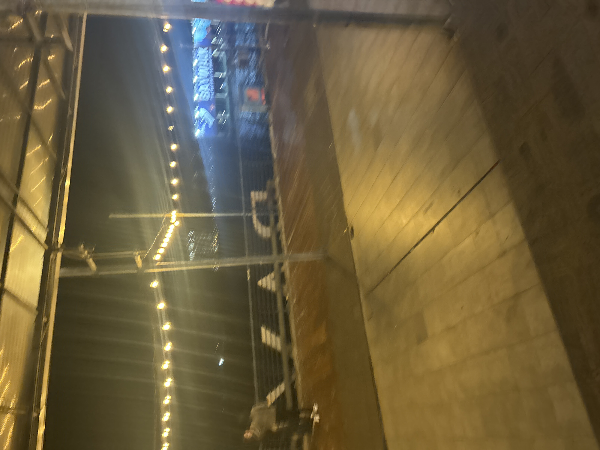 The image depicts a nighttime scene at Ancol Bay City, North Jakarta, Indonesia, specifically within the Baywalk area. A person, partially visible and out of focus on the left side, appears to be standing on a wooden boardwalk. String lights are illuminated overhead, casting a warm glow. In the background, a large blue banner with white lettering, reading "BAYWALK," is prominently displayed. The environment suggests an outdoor entertainment or dining district.