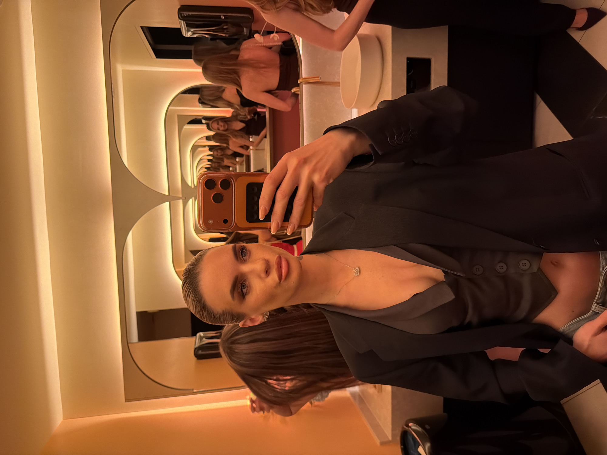 A fair-skinned woman with blonde hair is taking a selfie in what appears to be a bathroom. She's wearing a black blazer and a gray top, with a silver necklace visible. The phone case she's using is brown.
Behind her is a mirror that creates a repeating reflection effect, showing other women in the background. The bathroom has an orange-toned lighting and a modern design with a curved mirror and sink. The exact location in Berlin, Germany cannot be determined with certainty based on the image alone.