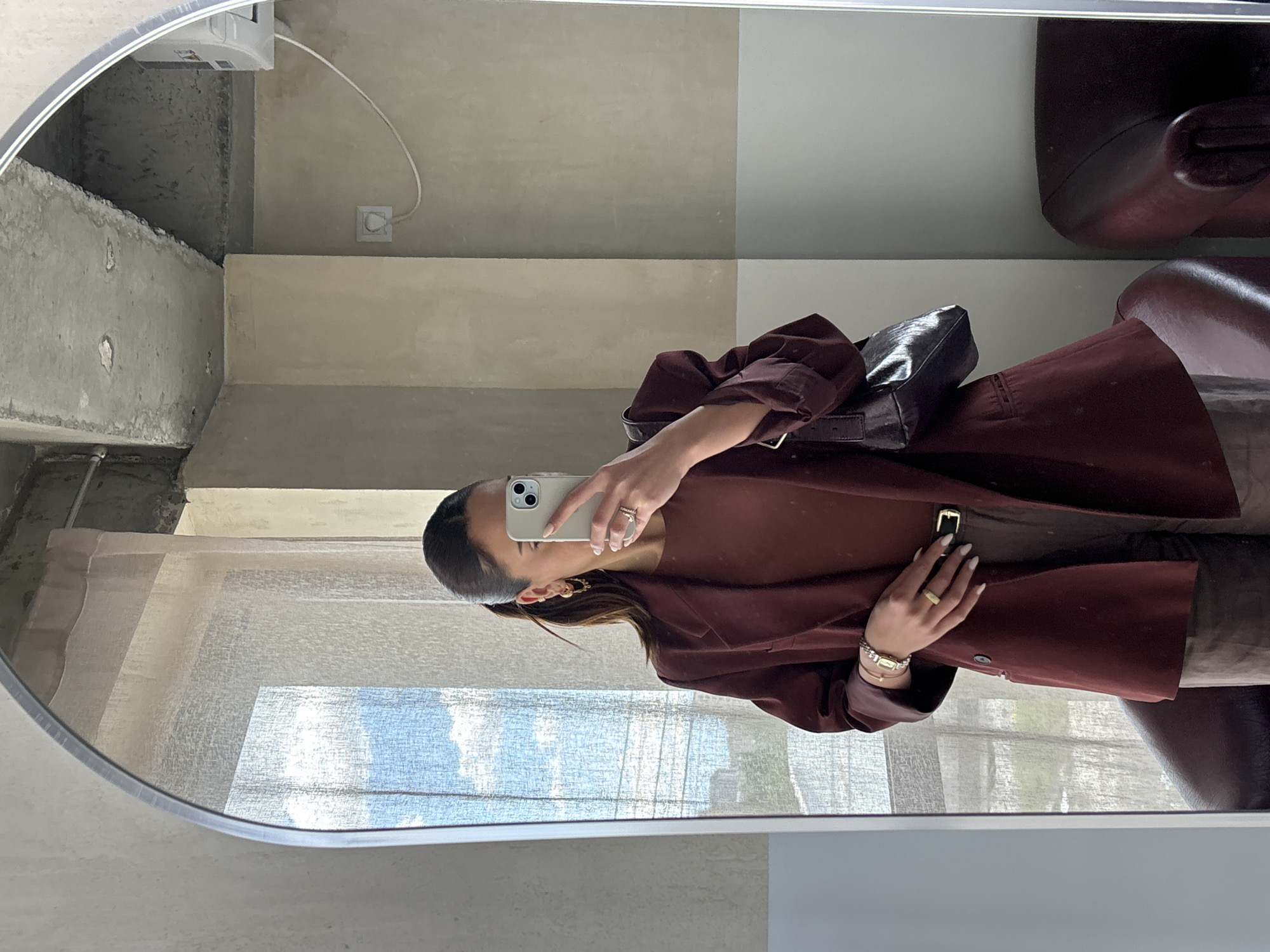 A person takes a mirror selfie, holding a smartphone to their face with their right hand. They are wearing a maroon colored blazer, a brown shirt underneath, and a dark handbag. The mirror is situated in front of a window with a light-colored blind, which is partially open, allowing light to enter the room. The wall behind them is divided into gray and light beige sections. In the reflection, pieces of furniture in a dark color are also visible. The exact location in Yerevan, Armenia cannot be determined from the image.