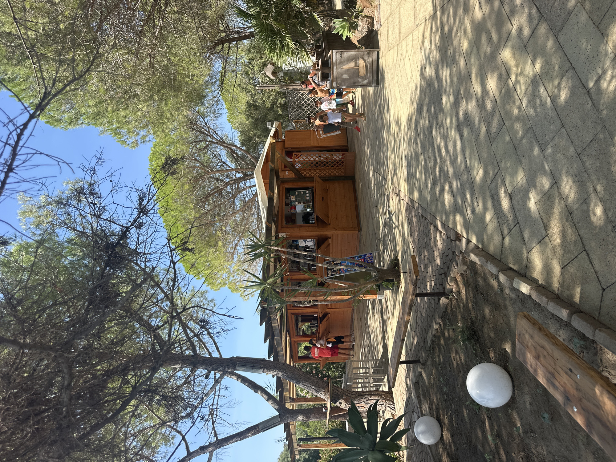 The image depicts an outdoor scene in what appears to be a park or recreational area, possibly in Biddeputzi/Villaputzu, Italy. In the center, there is a wooden kiosk or small building with a covered roof, serving as a bar or refreshment stand. Several people are visible around the kiosk. To the left, two individuals, one wearing a red shirt, stand near the counter. To the right, a group of children and at least one adult are interacting near the kiosk. One child, a young girl with a shovel, is walking towards the foreground. The ground is paved with light-colored rectangular stones, with some areas of dirt and landscaping, including trees and large white spherical decorative objects. Wooden benches are placed in the foreground and mid-ground. The environment is sunny, with dappled sunlight filtering through the trees onto the paved area.