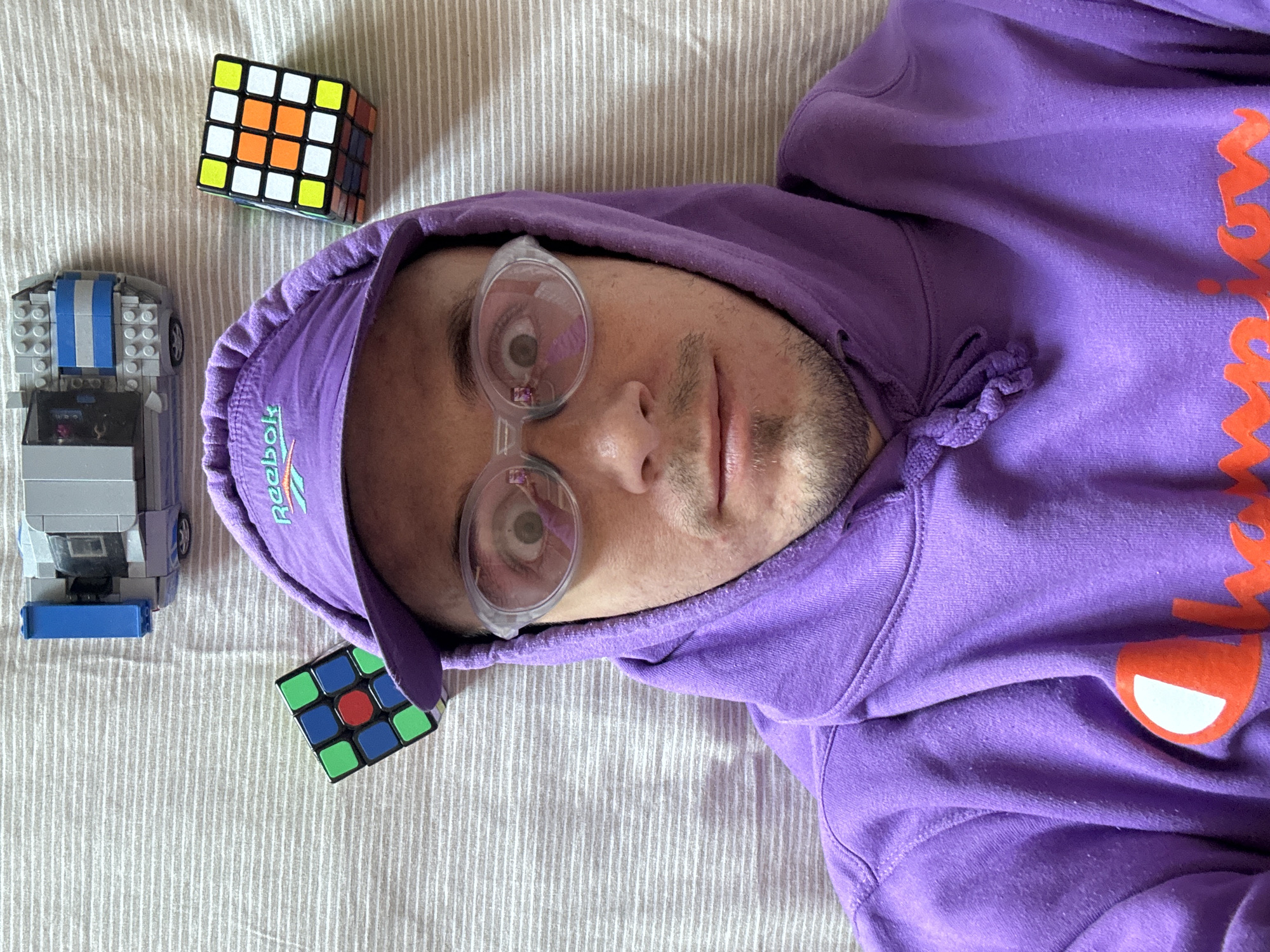The image is a close-up, overhead shot of a person lying on a striped white and grey surface, wearing a purple hoodie and a purple baseball cap with the Reebok logo. The person is wearing clear, oval-shaped glasses, which reflect a pink object. Two Rubik's cubes and a Lego car are placed on the surface around the person's head. There is no indication of the city of Đà Nẵng, Vietnam, or any specific urban environment. The scene appears to be indoors, possibly a bed or floor, with a neutral background.