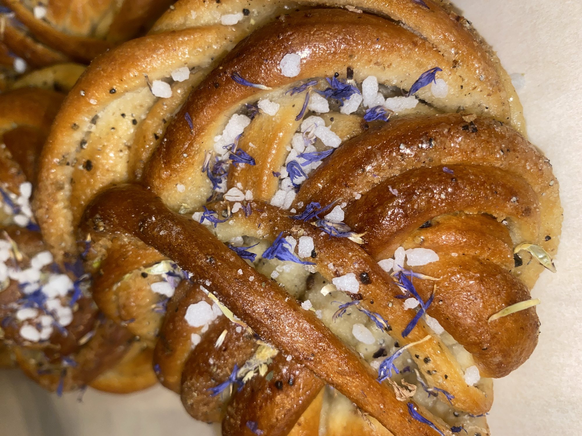 A close-up, overhead shot showcases a golden-brown, braided pastry, possibly a sweet bun or challah, adorned with coarse white sugar crystals and delicate blue flower petals, some of which are scattered amongst the braids. The pastry has a slightly glossy finish, indicative of an egg wash or glaze. There are also small dark specks, possibly poppy seeds or spices, visible on the surface. The pastry is resting on a light-colored, textured surface, likely paper or parchment. The lighting is warm, highlighting the textures and colors of the pastry. The image is taken from an angle that allows for a detailed view of the pastry's intricate braided pattern. There are no visible people or indications of a specific location within Warsaw, Poland.
