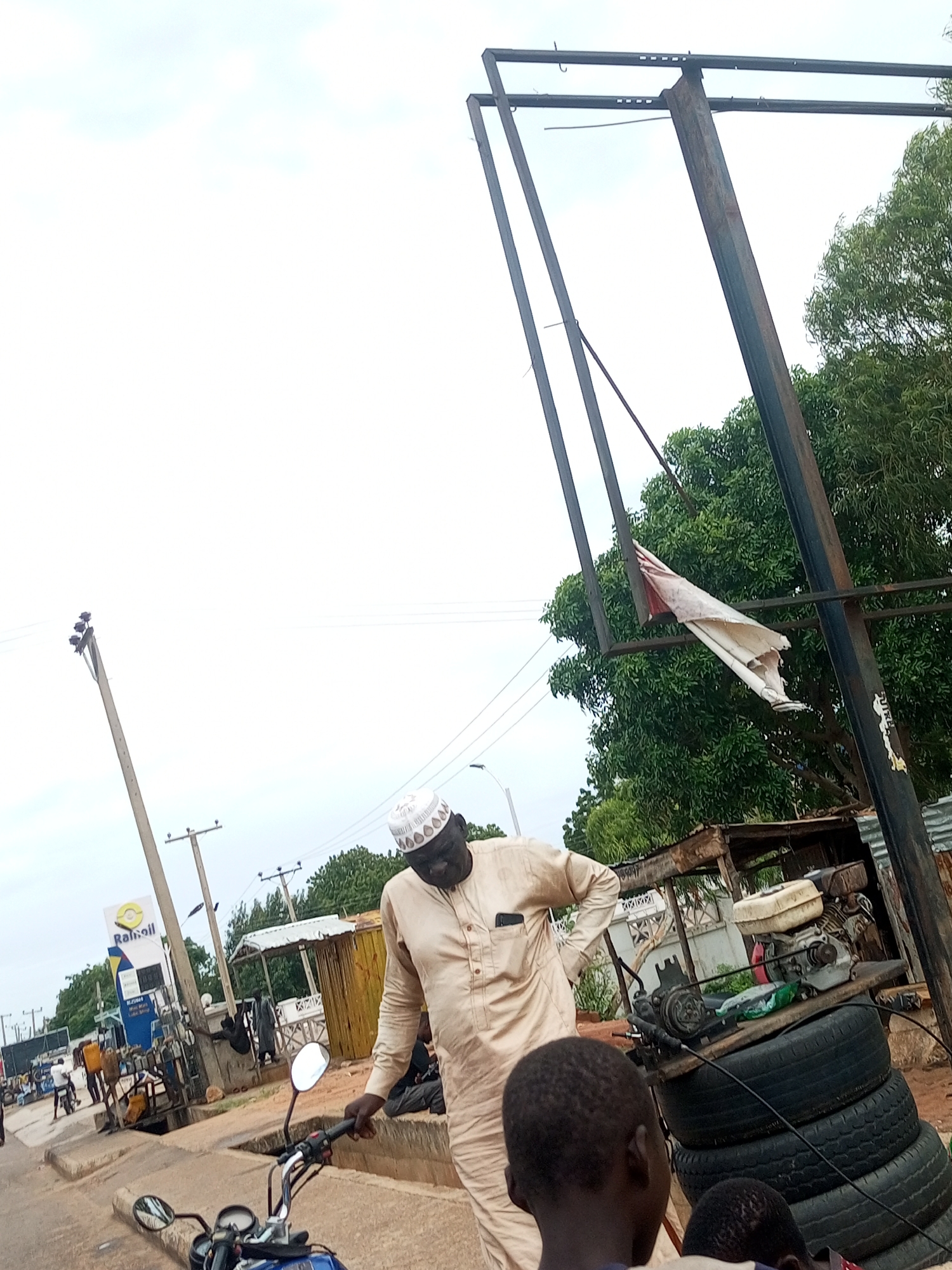 The image depicts an outdoor scene in Tempure, Nigeria. A man wearing a traditional beige kaftan and a white cap stands near a motorcycle, holding its handlebars. In the background, a gas station with a "Rainoil" sign is visible, along with other structures, trees, and power lines. A tall, metal frame structure with a tattered white cloth attached to it stands prominently. Several tires are stacked near a small engine. The sky is overcast.