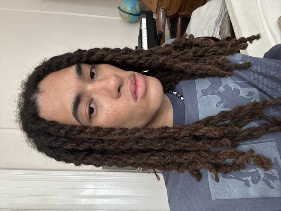 A forensic examination of the image reveals one individual positioned centrally in a static portrait. The person has an apparent mixed or ambiguous ethnicity, light brown skin with visible freckles, and long, dark brown dreadlocks that extend past the shoulders and over the chest. They have dark brown eyes and maintain a direct gaze towards the camera with a neutral facial expression, lips slightly parted. The individual wears a dark grey t-shirt featuring a faded light grey graphic design on the front. A beaded necklace, comprising small white, black, and brown cylindrical beads, is visible around the neck.
The scene is an interior room in London, United Kingdom. To the subject's left, a white, vertically paneled door or wall section is visible. To the subject's right, a black electronic keyboard or digital piano is positioned, with a light blue globe visible above and behind it. A dark wooden chair with a curved backrest is partially discernible below the keyboard. Further to the right, a light-colored wooden furniture piece and a rumpled white sheet or textile, consistent with bedding, are present. The walls of the room appear light-colored.