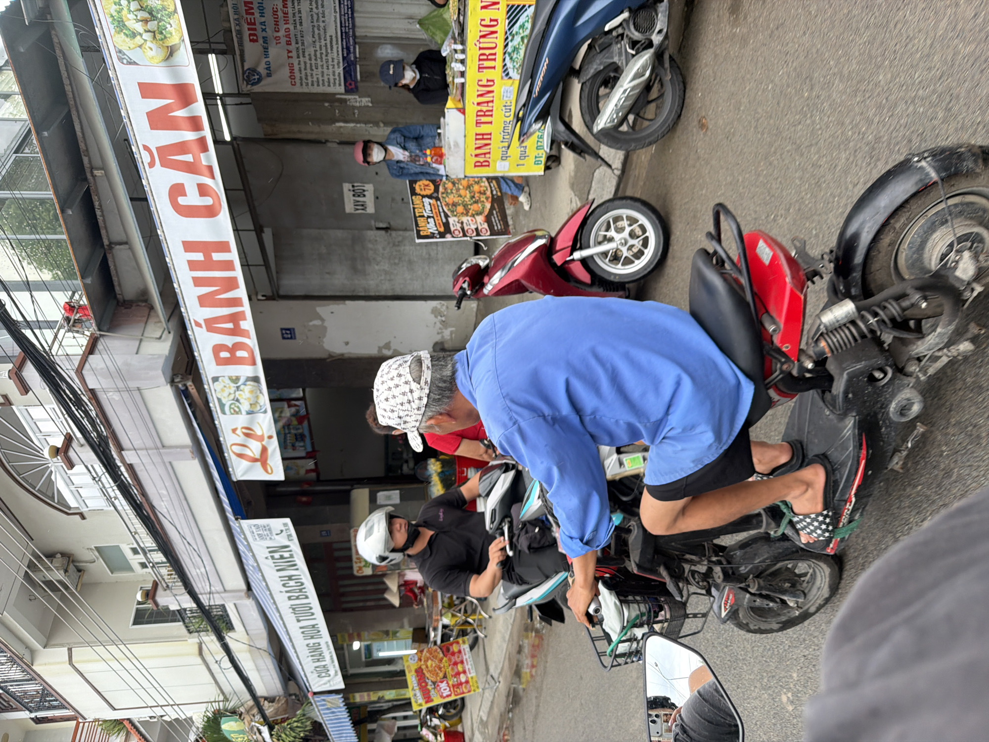 The image depicts a street scene in Nha Trang, Vietnam, featuring a row of food stalls and shops. In the foreground, a person wearing a blue shirt and black shorts rides a red electric scooter. Behind them, another person in a black polo shirt and black pants, wearing a white helmet and a black face mask, rides a white and blue electric scooter. Several other individuals are visible at the food stalls, including a woman in a denim jacket and pink helmet, and another in a black shirt and blue cap, both wearing face masks. Scooters and motorcycles are parked along the street, and various signs advertise food items, including "BÁNH CĂN" and "BÁNH TRÁNG TRỨNG NGON". The environment suggests a bustling urban setting with overhead wires and shop awnings contributing to the streetscape.