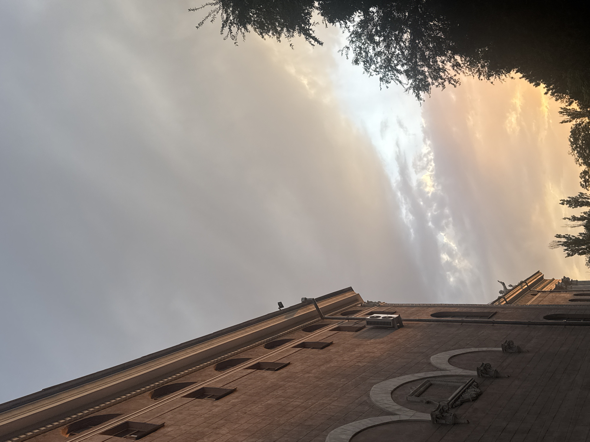 The image shows the facade of a brick building with arched windows, an air conditioning unit, and decorative architectural elements. The building is seen from a low angle, with the sky taking up most of the frame. The sky displays a sunset with clouds in various shades of grey, orange, and yellow. Silhouettes of trees are visible on the right side of the image, and some tall, thin trees are also visible in the distance at the bottom. There are no people or animals present in the scene. The location is identified as Bishkek City, Kyrgyzstan.