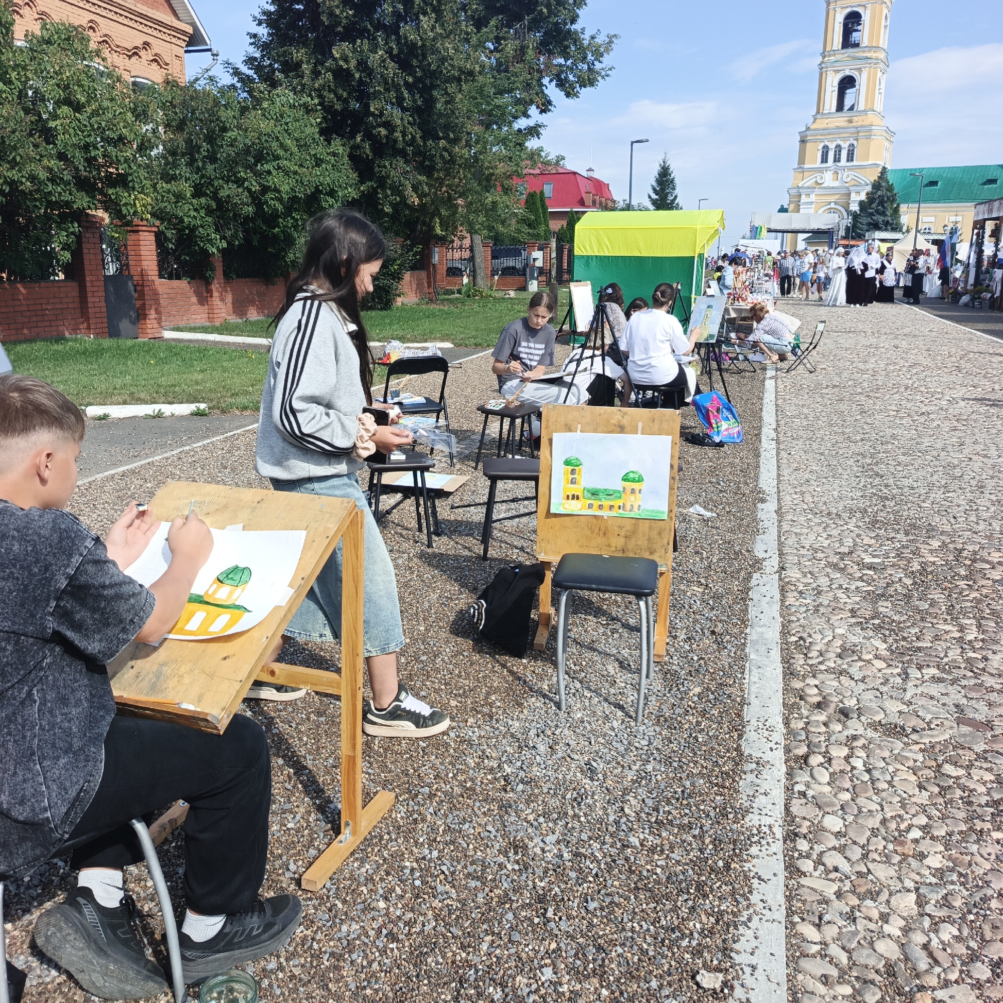 In Nikolo-Beryozovka, Russia, on a sunny day, an outdoor art class is taking place on a cobblestone path. Several young artists are seated on stools, working on their paintings at wooden easels. In the foreground, a boy in a dark gray t-shirt and black pants is actively painting a picture of a building with green domes. To his right, a girl in a gray sweater with black stripes and blue jeans is also engaged in painting. Behind them, a yellow and green tent is set up, and further in the background, a tall, ornate yellow bell tower of a church stands against a clear blue sky. Other people, including adults and children, are visible in the distance, suggesting a public event or gathering. The environment is characterized by a mix of green grass, trees, and the textured cobblestone ground.