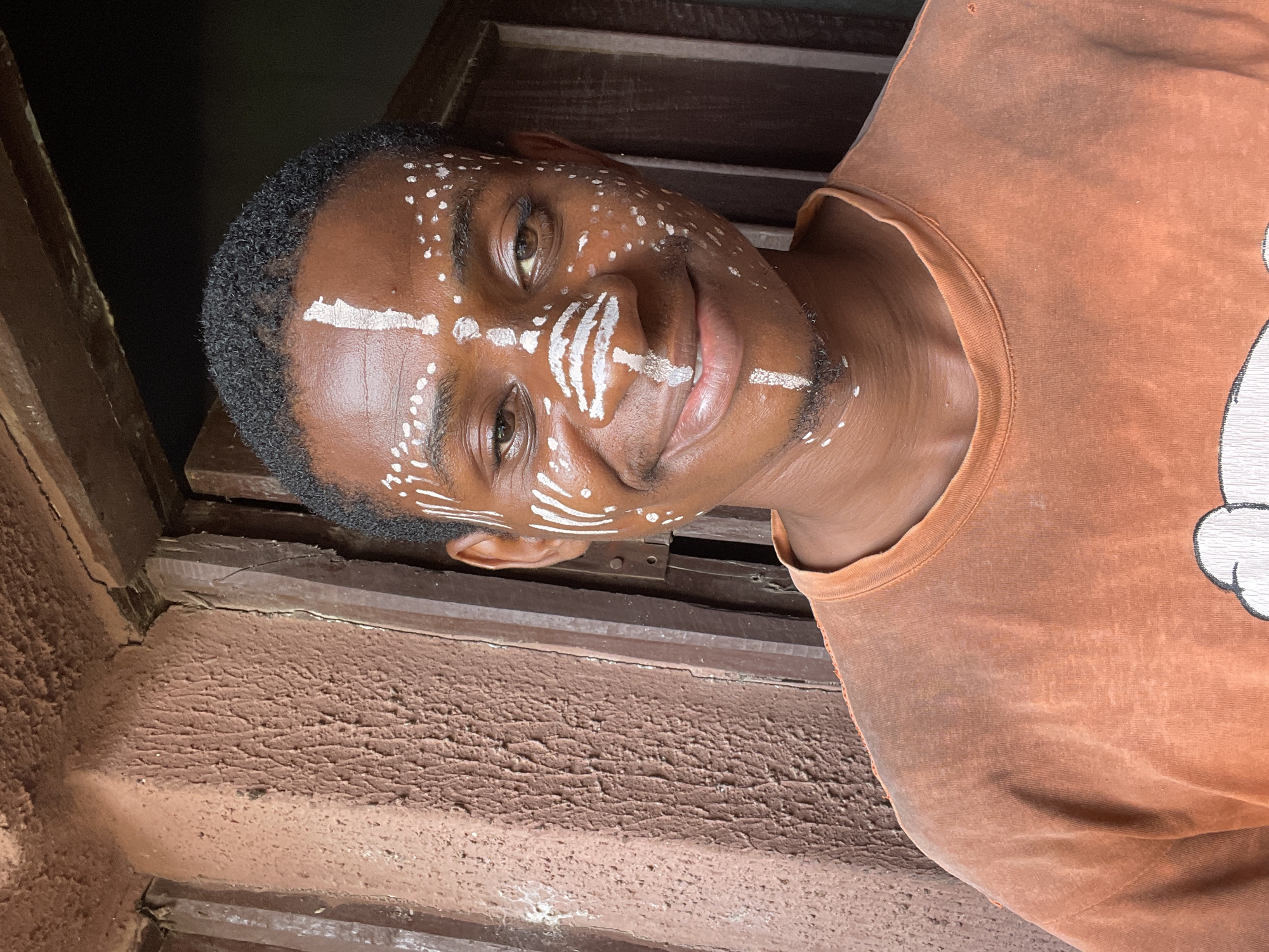 A young man with dark skin and short, dark, curly hair looks directly at the camera with a slight smile. He is adorned with intricate white facial markings, characteristic of traditional African body art. These markings include small white dots scattered across his forehead and cheeks, a prominent cross-like design on his forehead, multiple short horizontal lines extending across the bridge of his nose and beneath his eyes, and a single horizontal line on his chin. He wears an orange or rust-colored t-shirt, with a partial white graphic design visible on the right side of his chest, resembling part of a cartoon character or symbol. The image appears to be taken indoors or in a semi-enclosed space, with a background featuring rustic, reddish-brown textured walls or surfaces, possibly made of mud or rough plaster, and dark wooden architectural elements like beams or window frames. The overall setting appears simple and traditional, aligning with the cultural context of Nkpor, Nigeria. No discernible text is visible other than the partial graphic on his shirt.