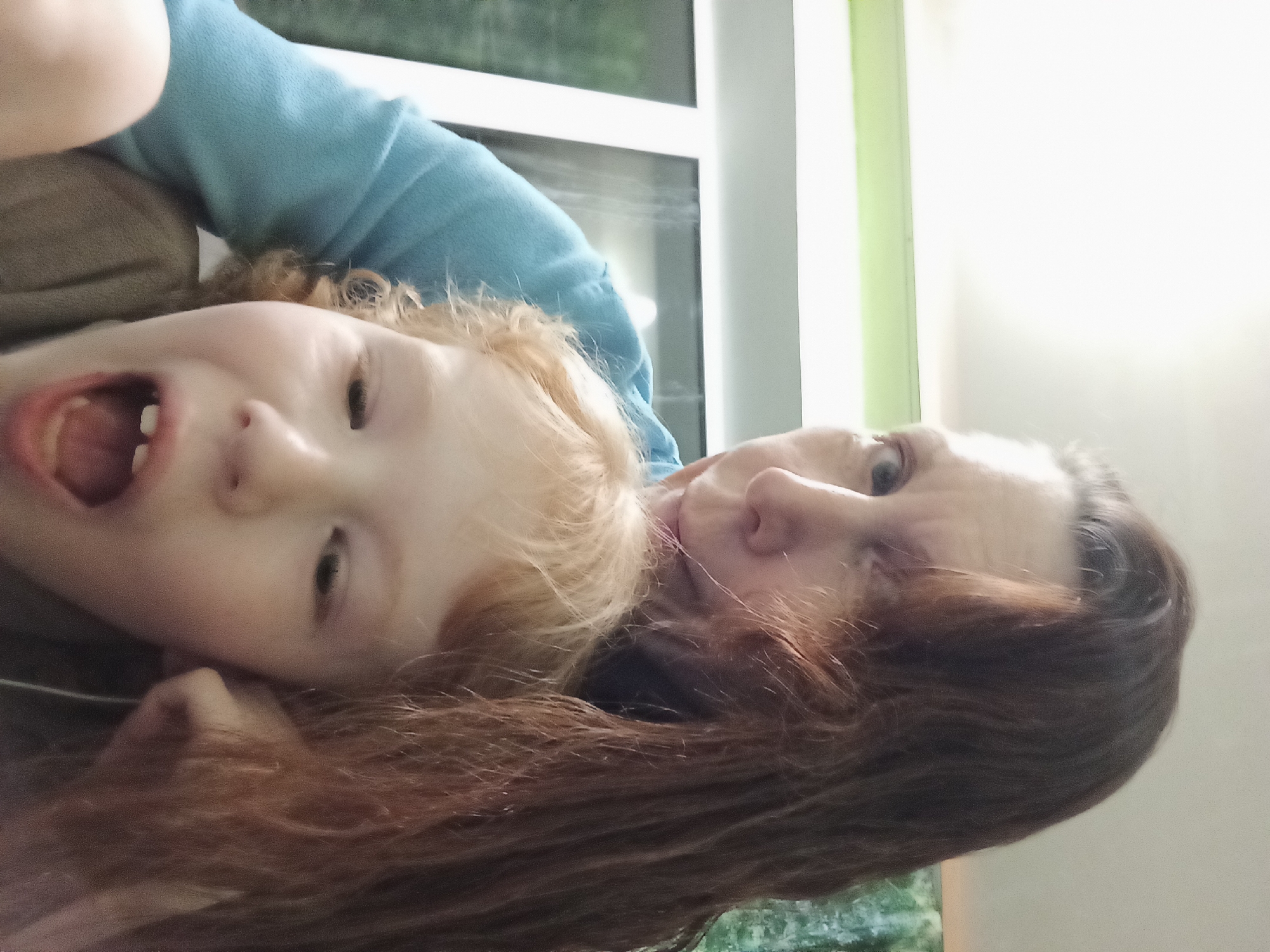 The image shows a woman with long, dark hair and a child with light-colored hair. The woman is wearing a blue top and is looking towards the camera. The child is in front of the woman and has their mouth open, as if speaking or shouting. The background appears to be indoors, with a window visible. The image quality is grainy and slightly blurry. There is no identifiable information in the image that would indicate its location in Blagoveshchensk, Russia.